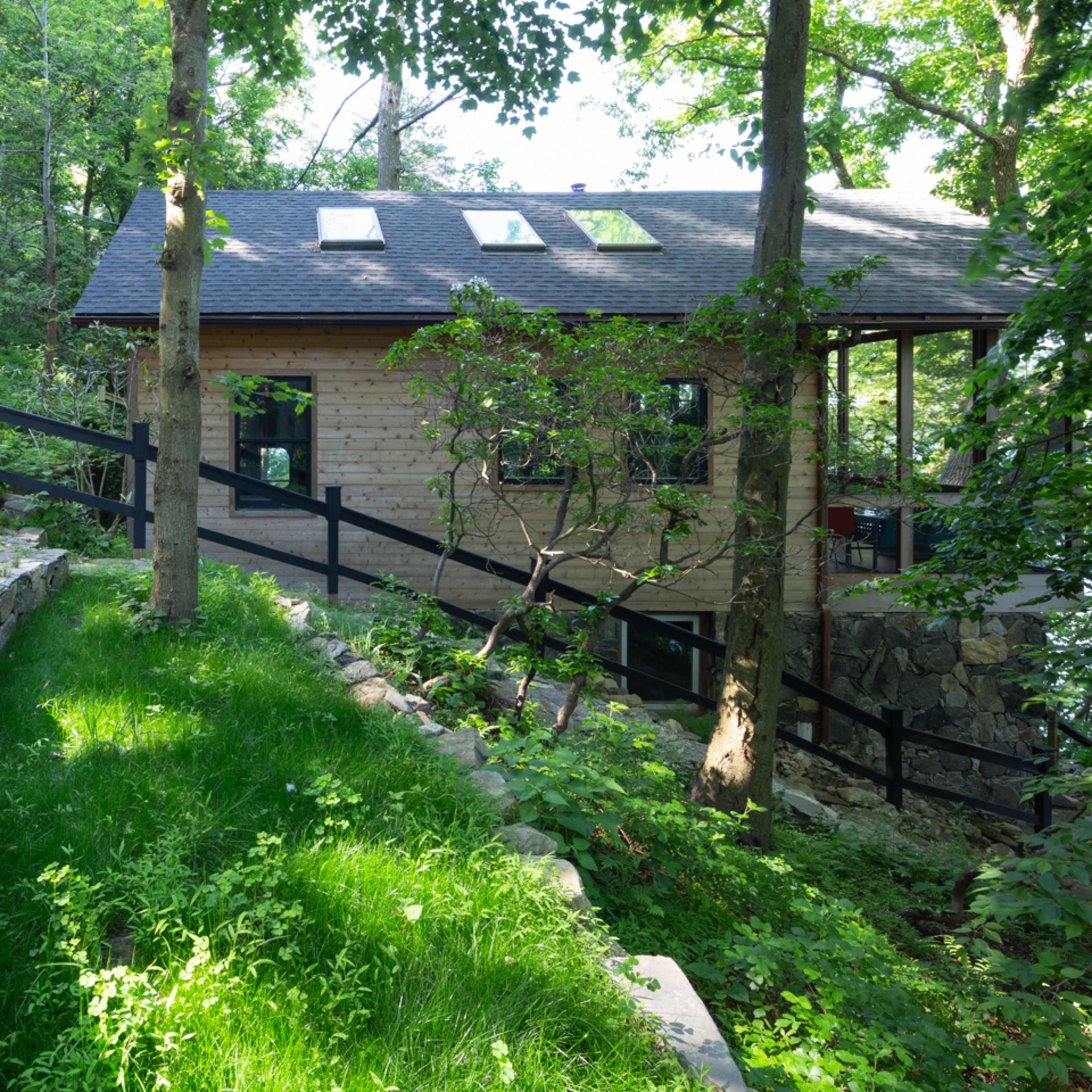 A wooden house with large windows is surrounded by trees and greenery next to a path leading down to a water source.