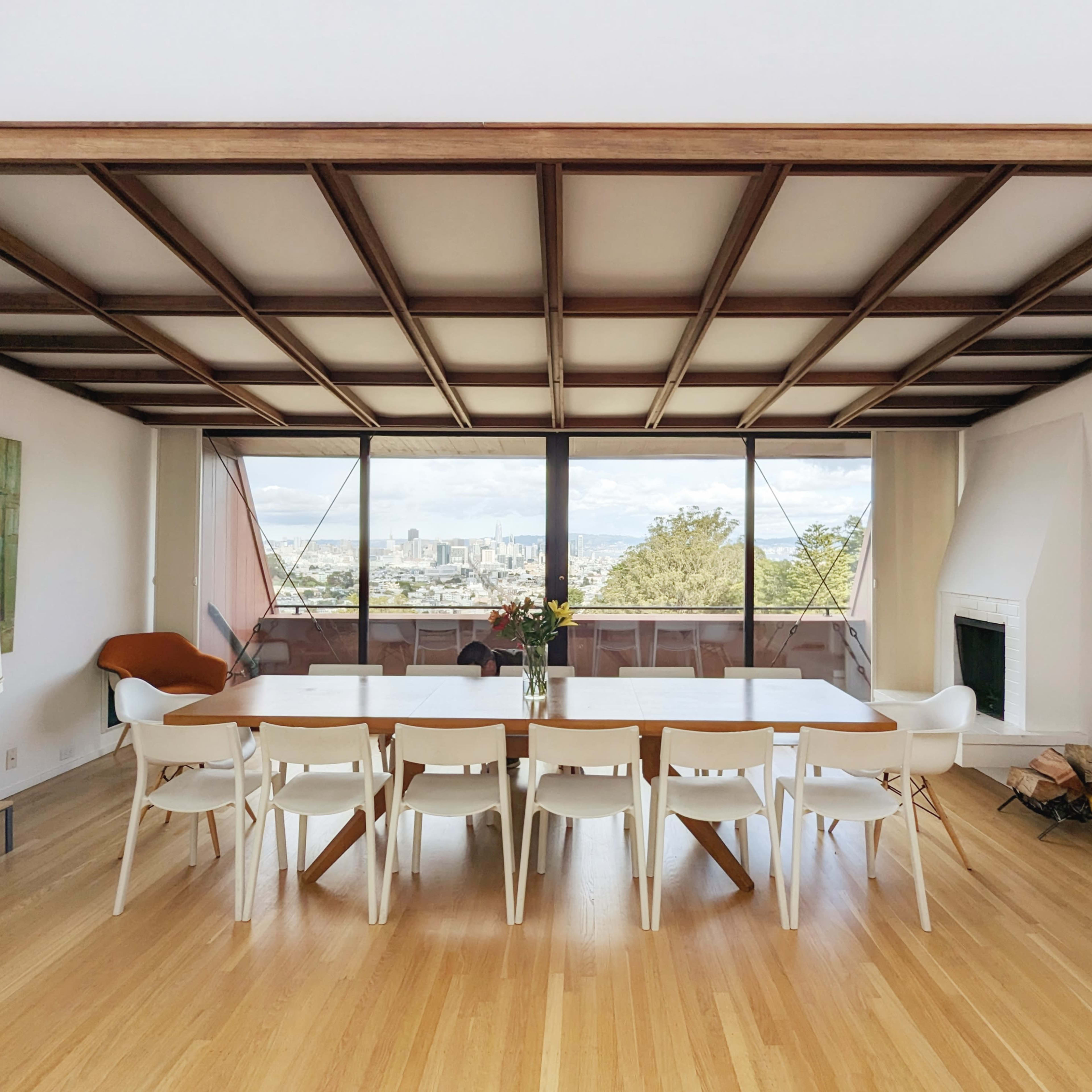 A spacious dining area features a long table surrounded by white chairs, with large glass windows offering a view of the city and mountains beyond.