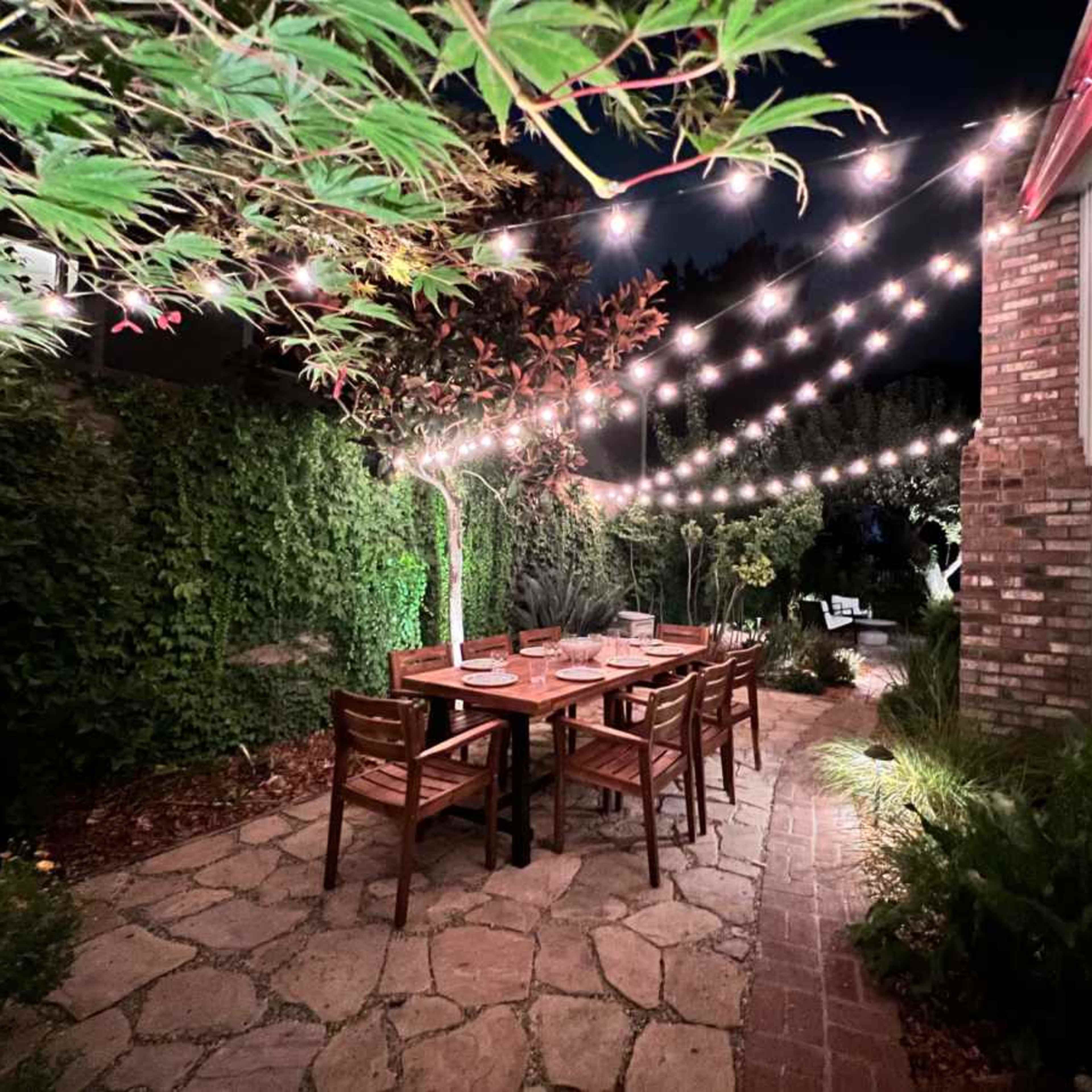 A stone pathway leading to a wooden dining table surrounded by chairs, with string lights illuminating the area at night.