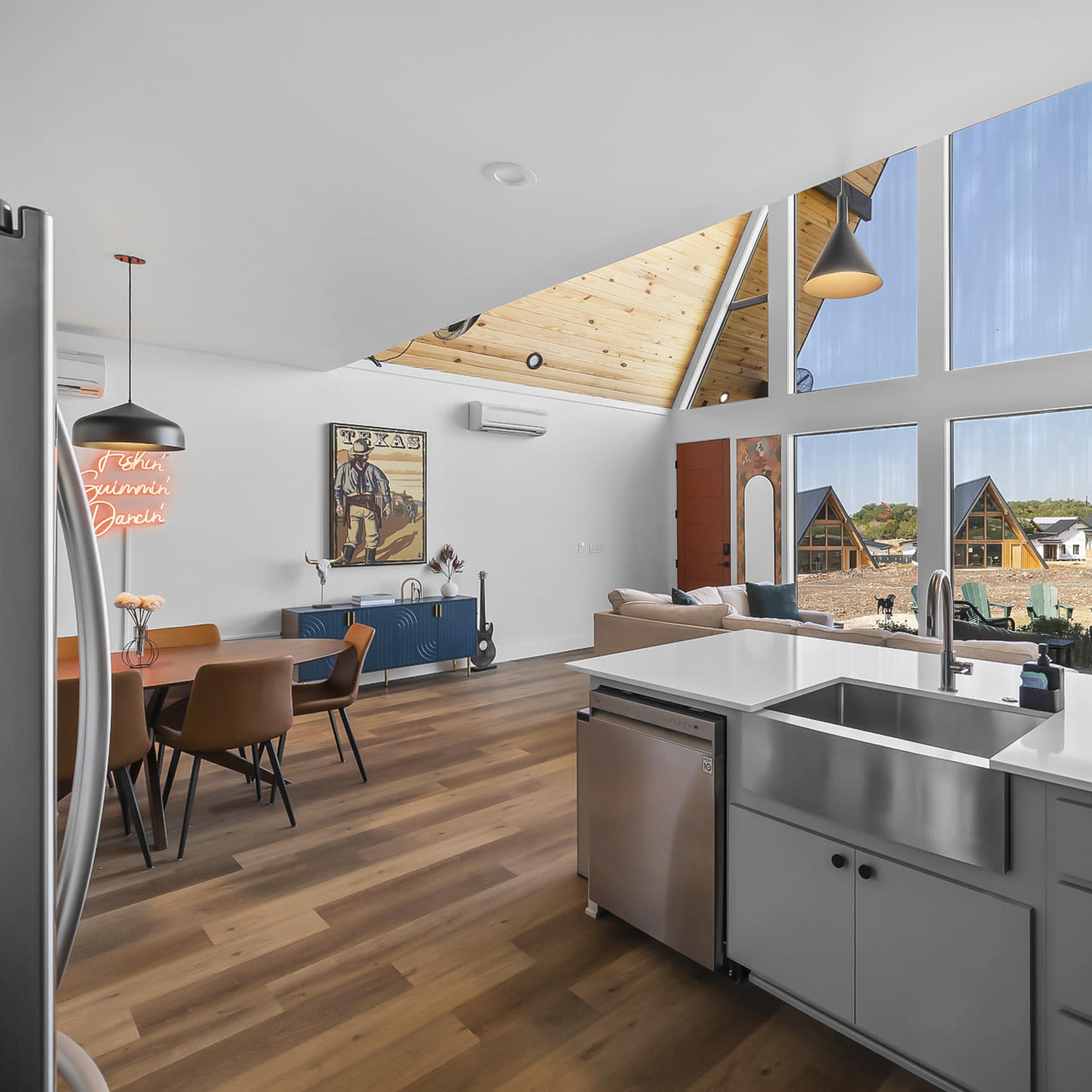 A modern kitchen and living area with large windows, wooden ceilings, and a dining space featuring a colorful artwork on the wall.