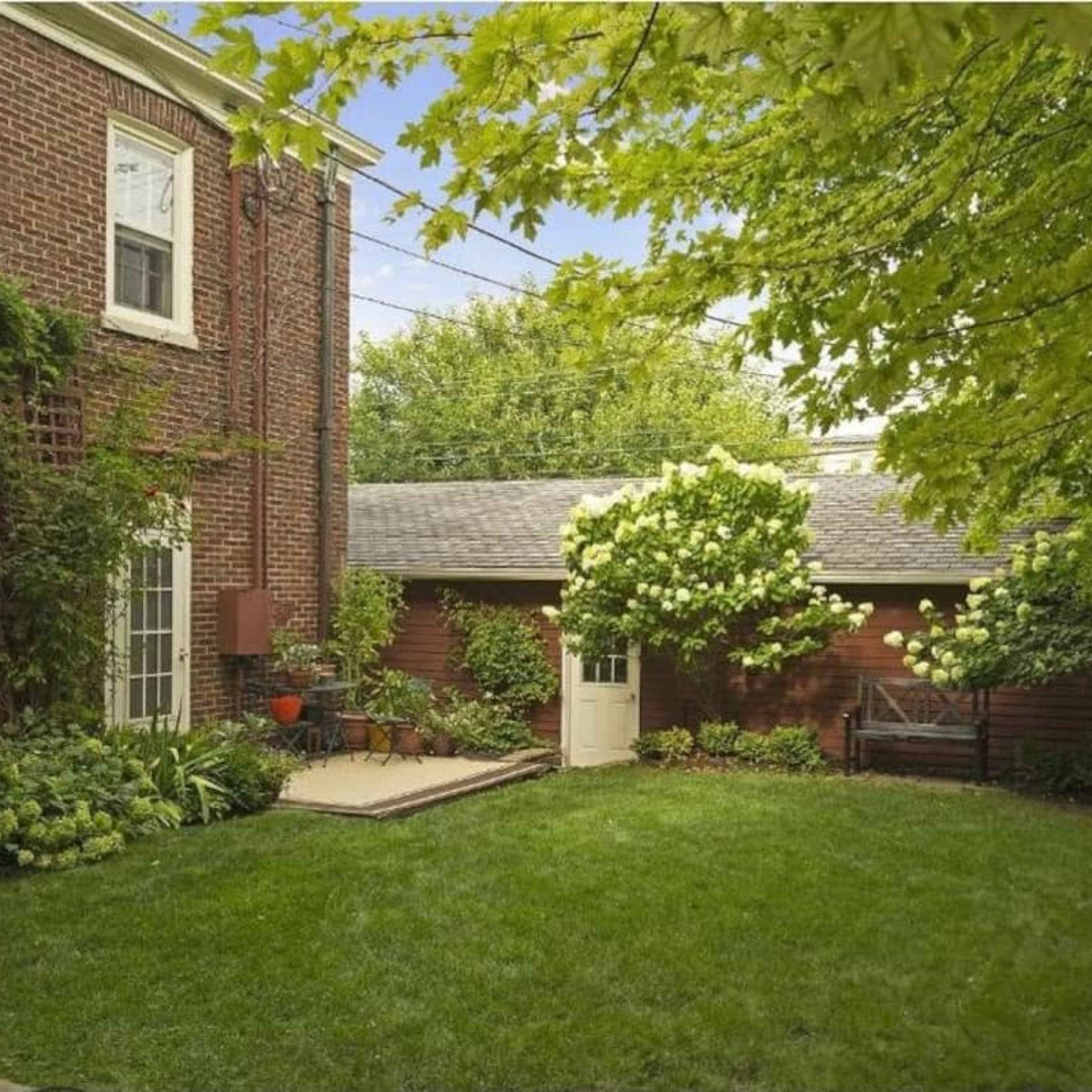 A backyard with a green lawn, surrounded by brick walls, trees, and shrubs, featuring a small patio area and a shed.