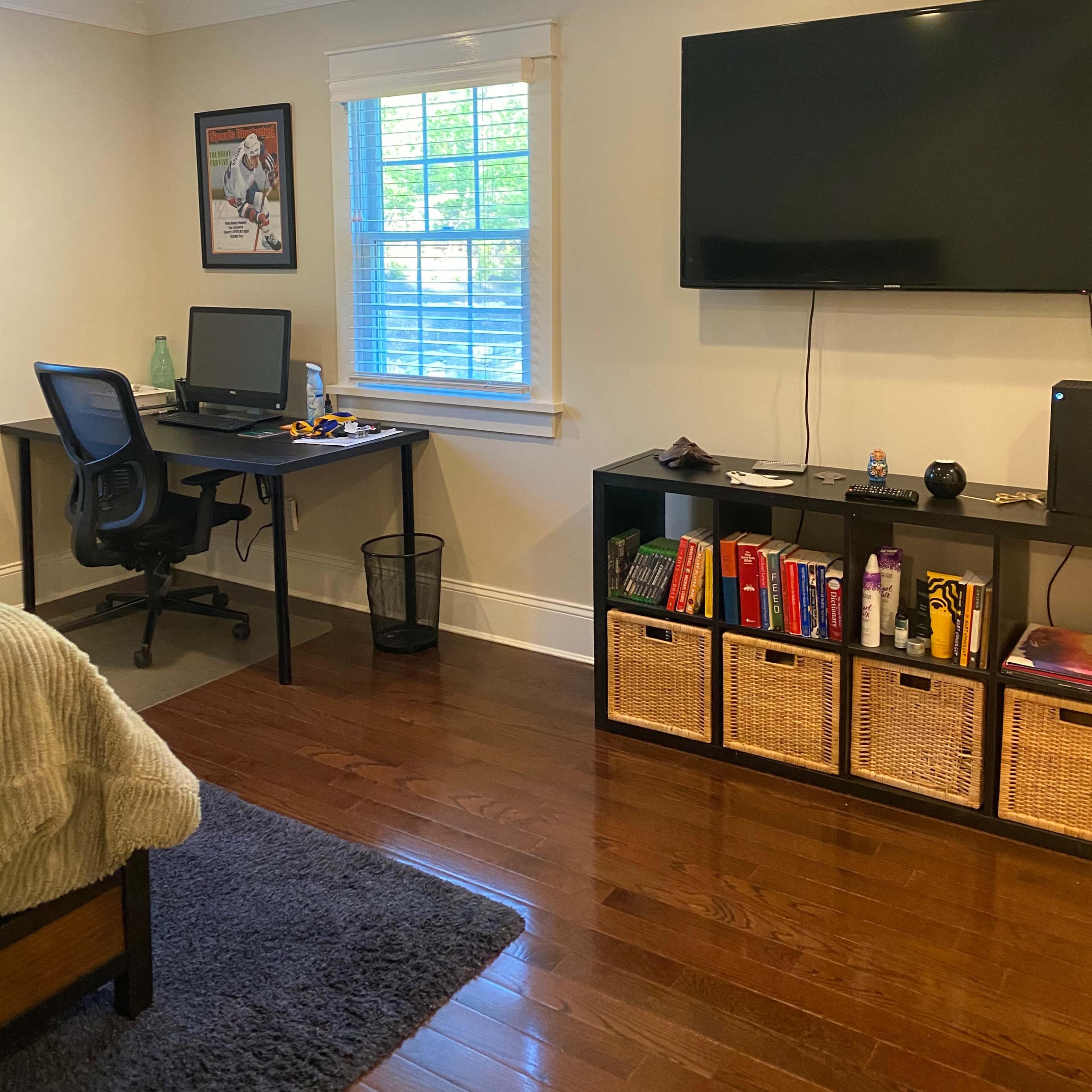 A home office area features a desk with a computer, a comfortable chair, and shelves with storage baskets, adjacent to a bed and a wall-mounted television.