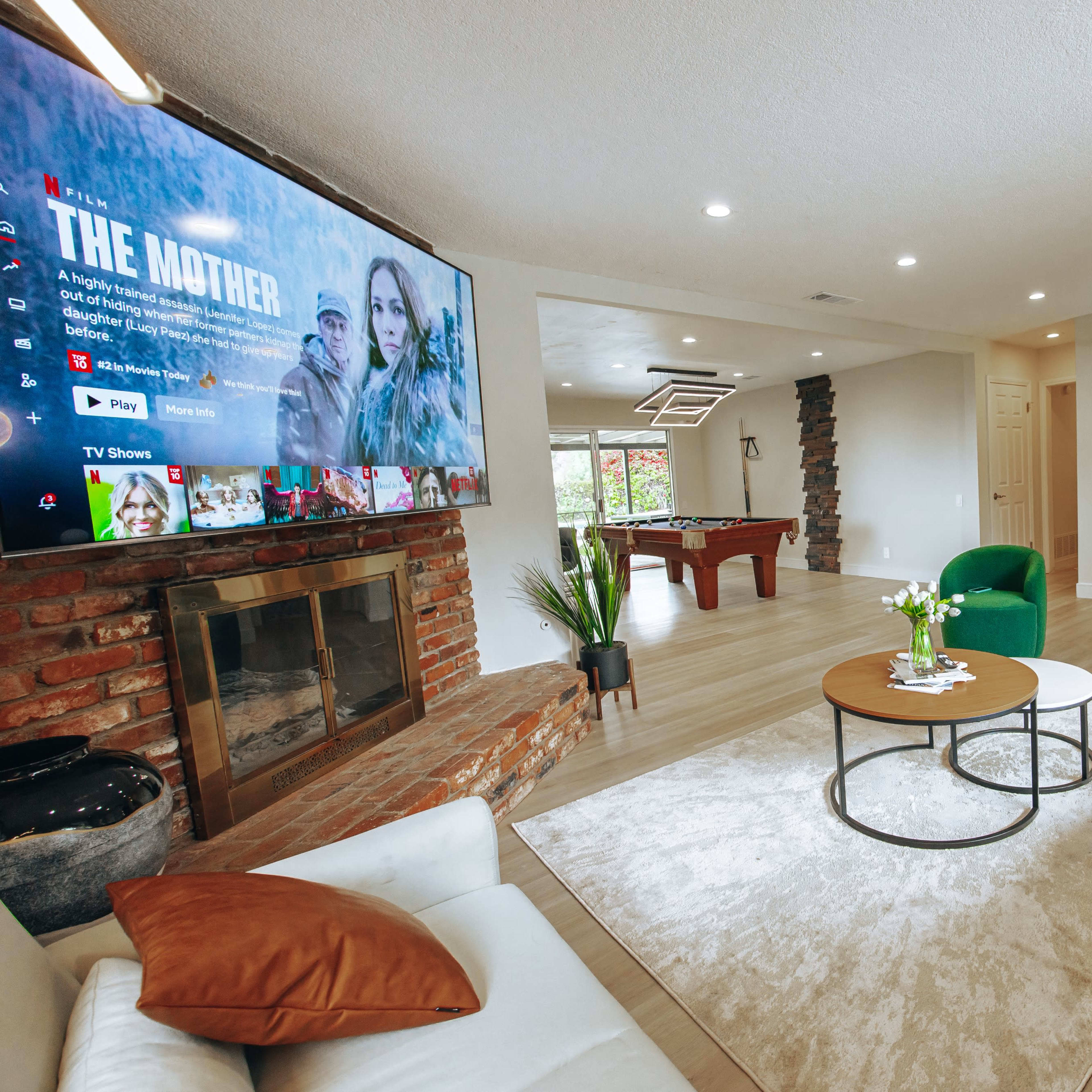 A modern living room featuring a large television above a fireplace, a pool table in the background, and comfortable seating arranged around a circular coffee table.