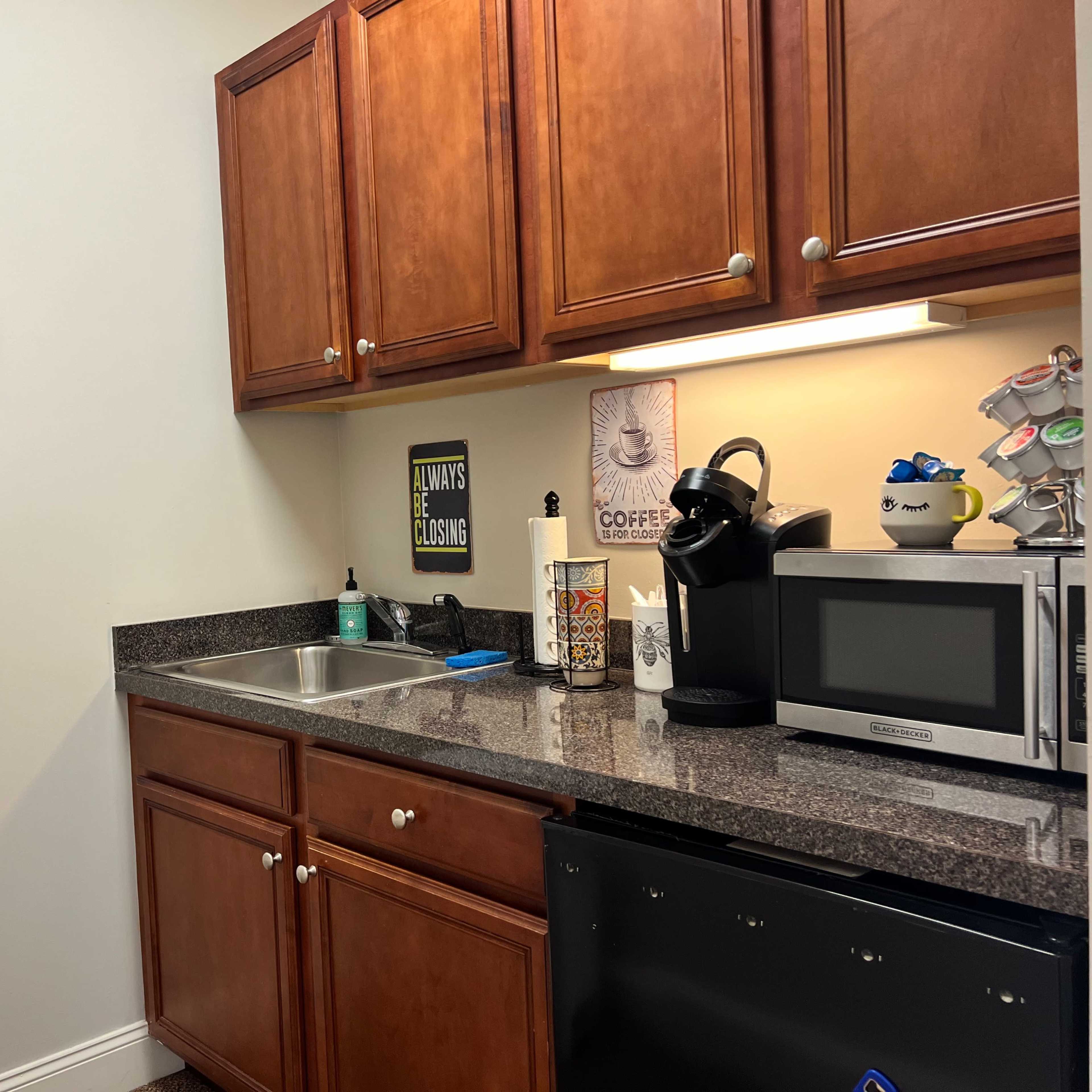 A kitchen corner features wooden cabinets, a clock on the wall, a coffee machine, a microwave, and a countertop with a sink.