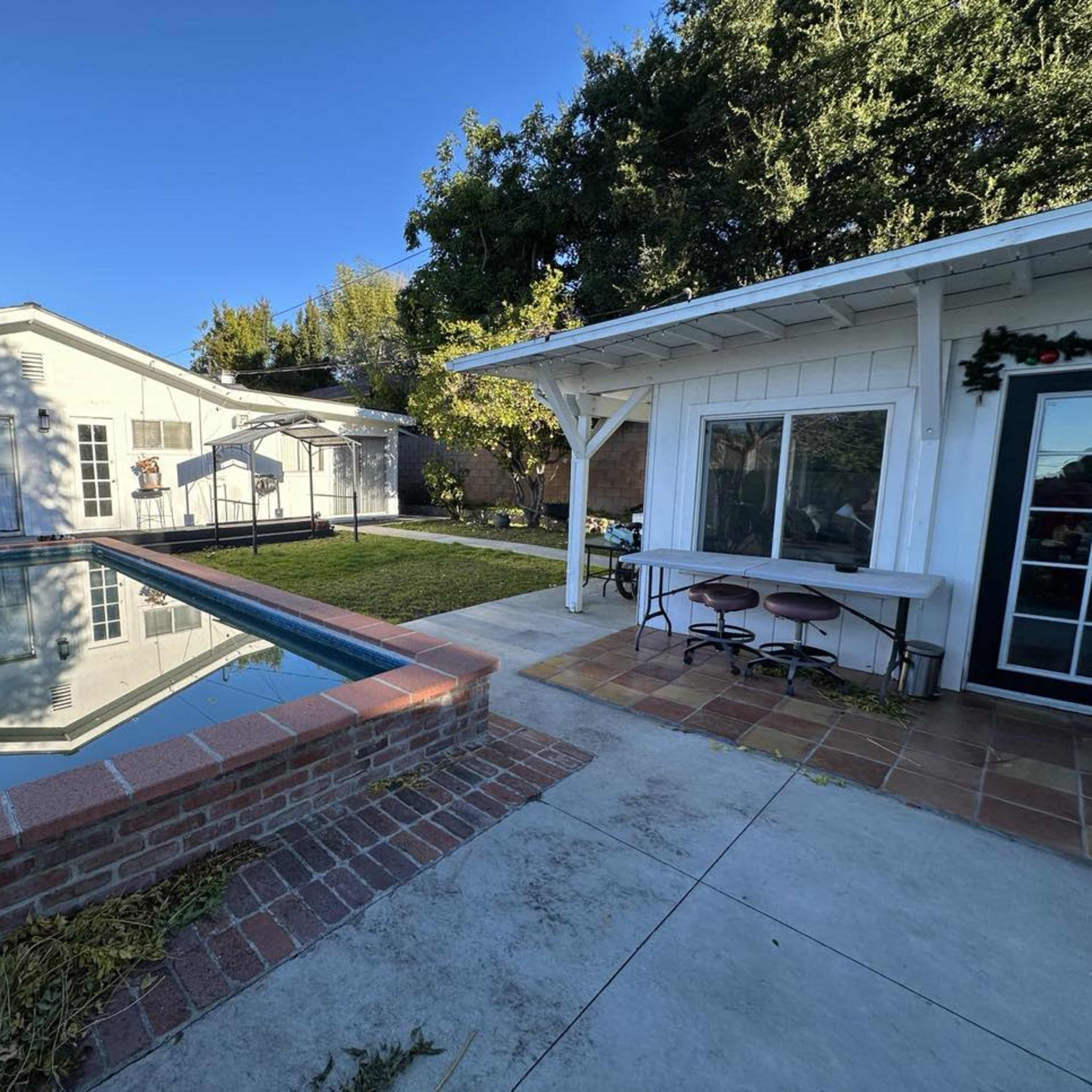 A backyard with a swimming pool surrounded by a patio, a small grass area, and a white house with a covered porch.