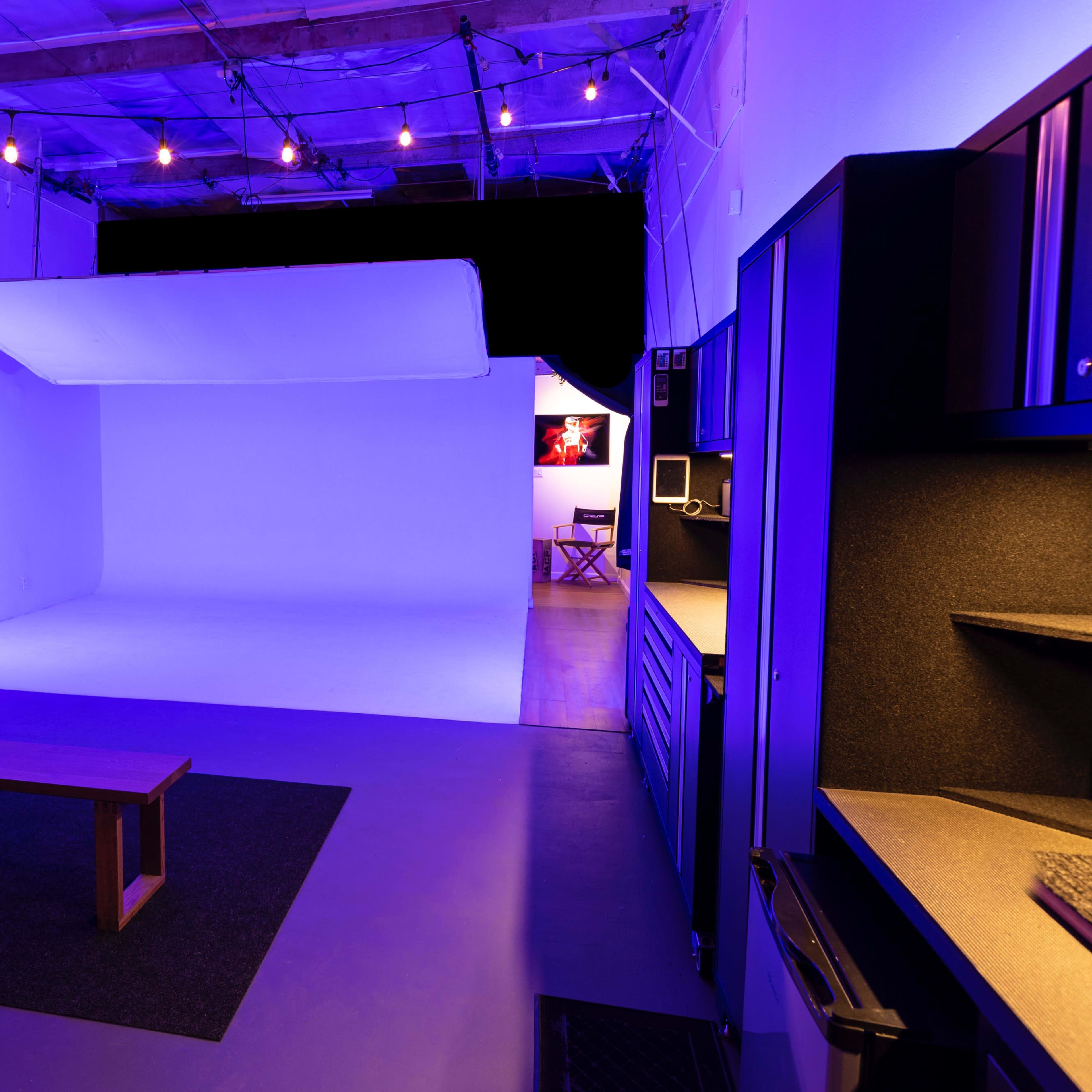 A lit studio space featuring a seamless white backdrop, a wooden bench, and a kitchenette area with overhead lights.