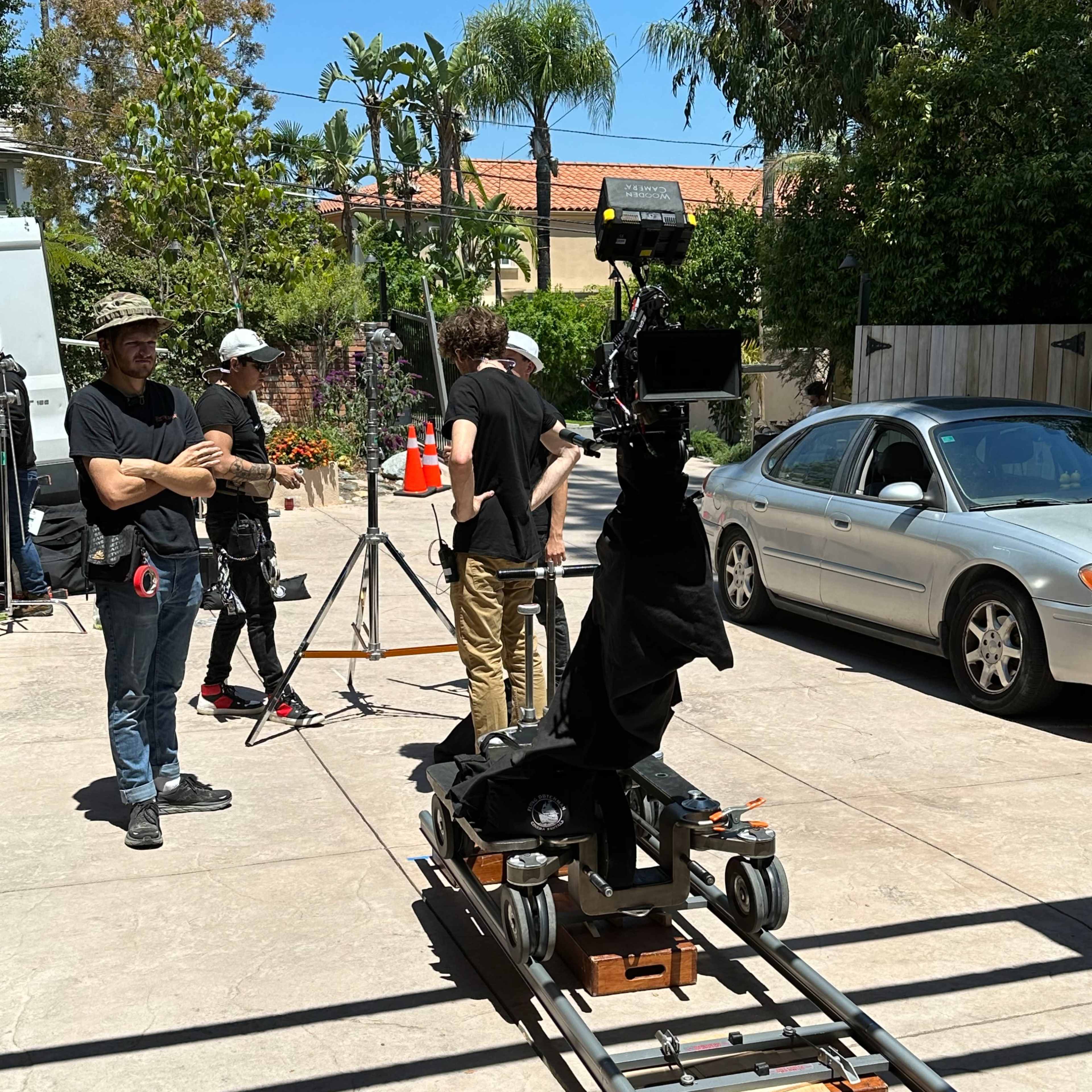 A film crew prepares a camera setup on a dolly track in a driveway with palm trees and parked vehicles in the background.