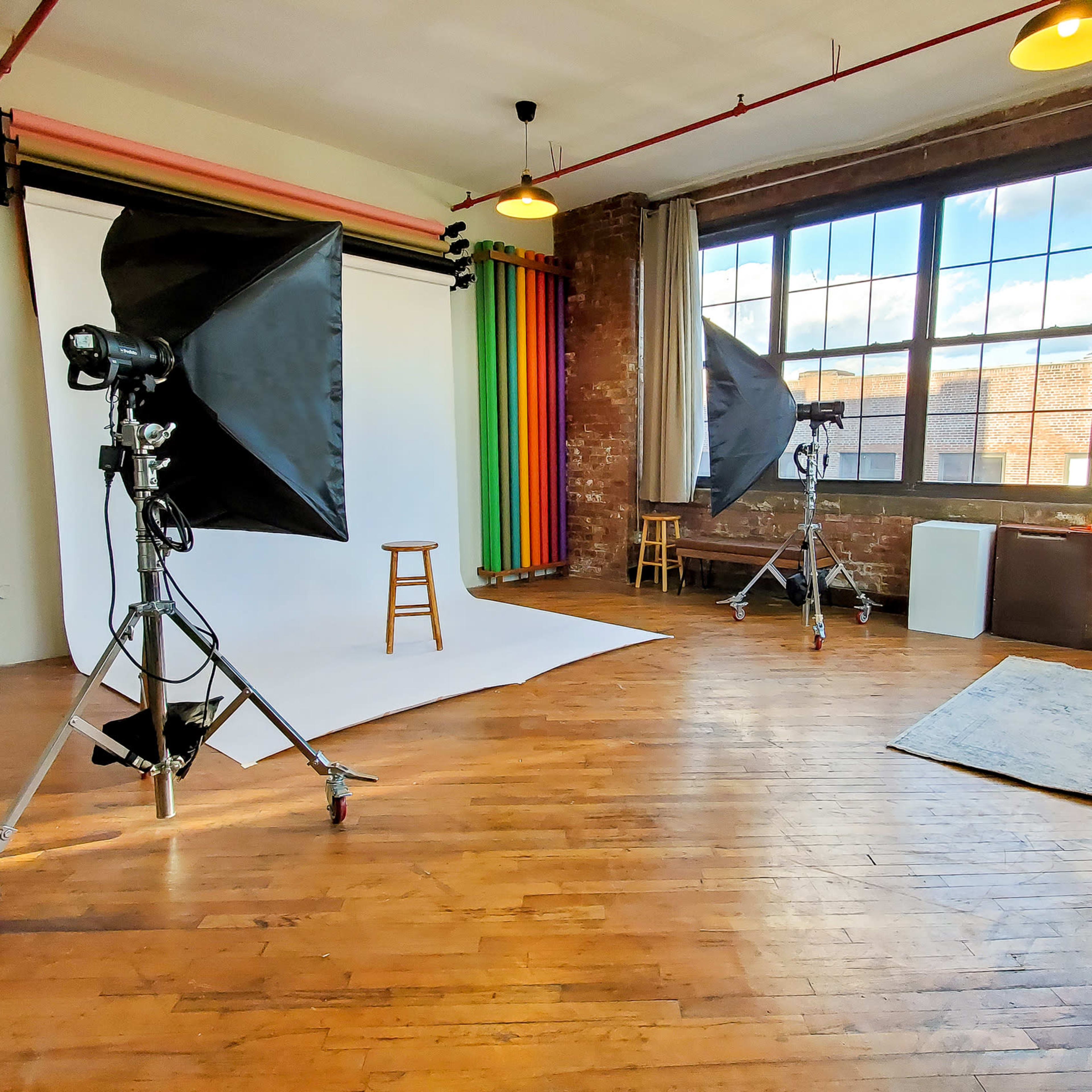 A photography studio with a white backdrop, lighting equipment, a stool, and a large window illuminated by natural light.