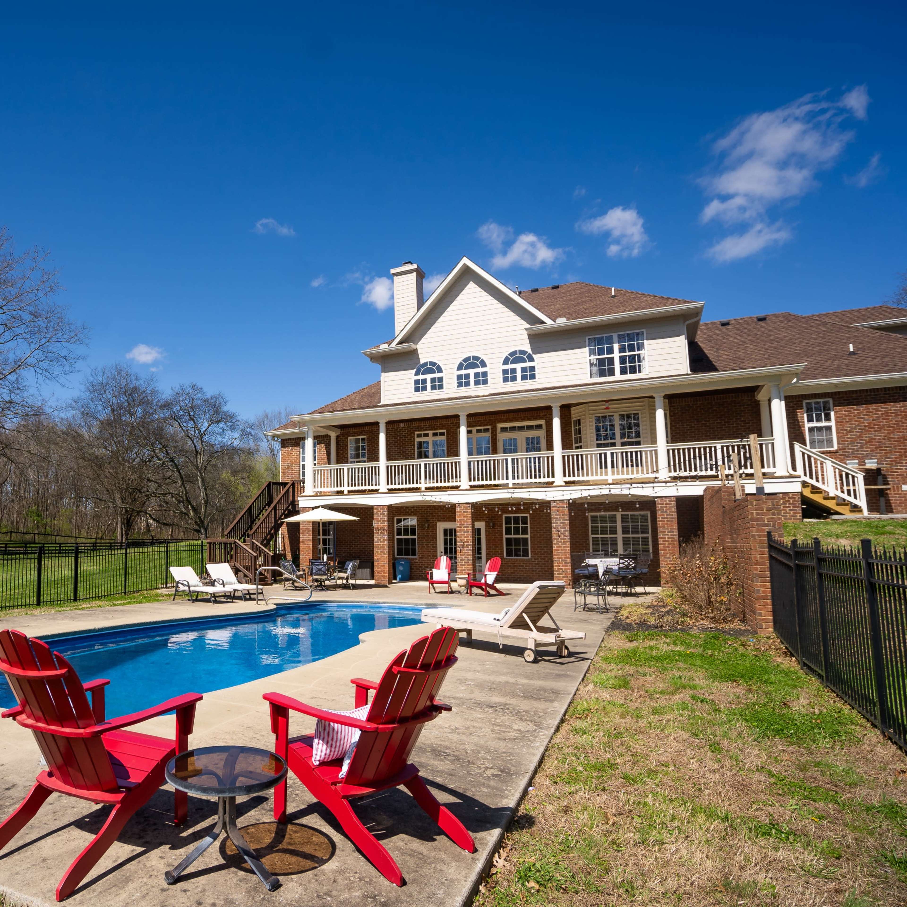 A backyard features a swimming pool surrounded by lounge chairs and a patio, with a spacious two-story house in the background.