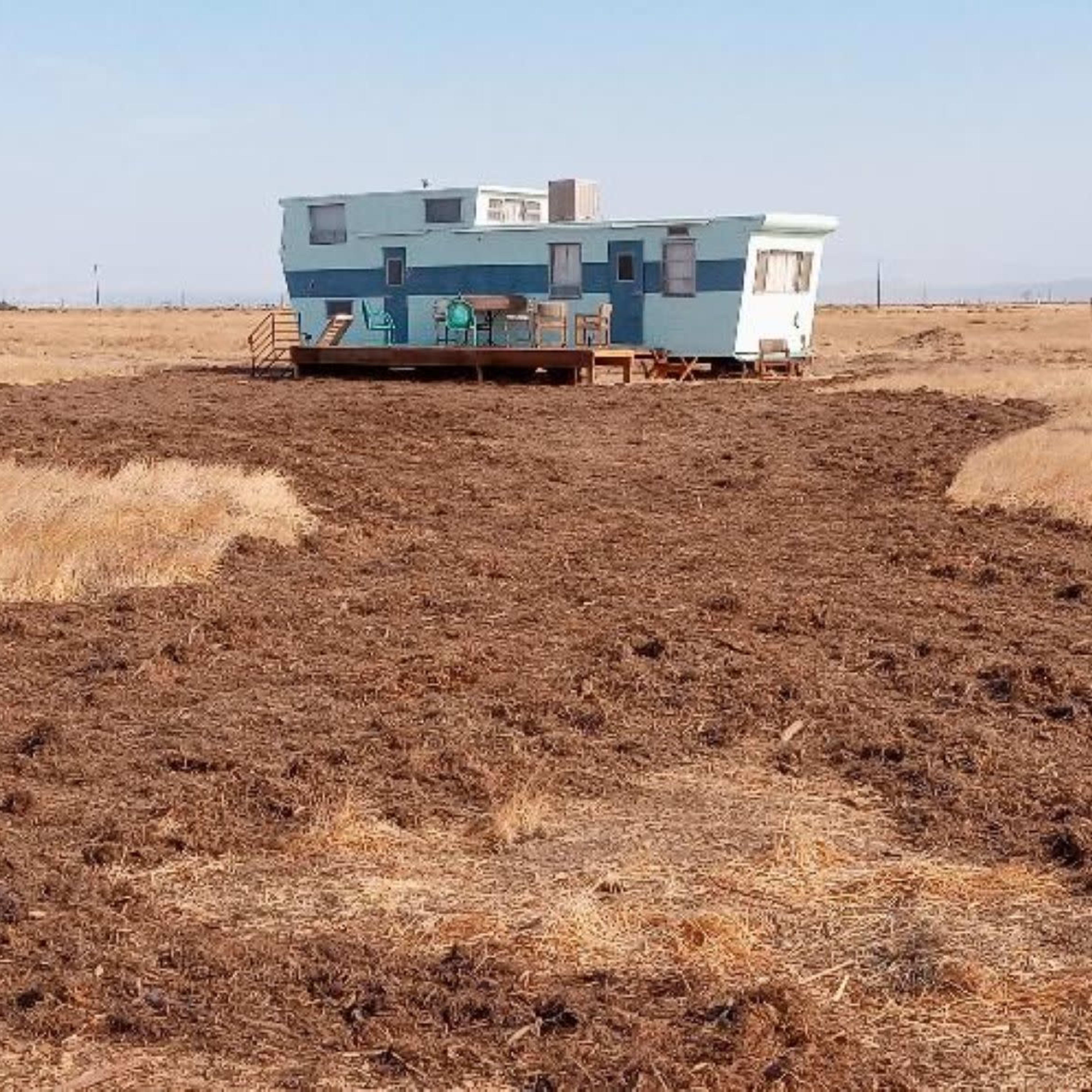 A blue trailer sits on a patch of dry grass surrounded by a large area of freshly turned soil.