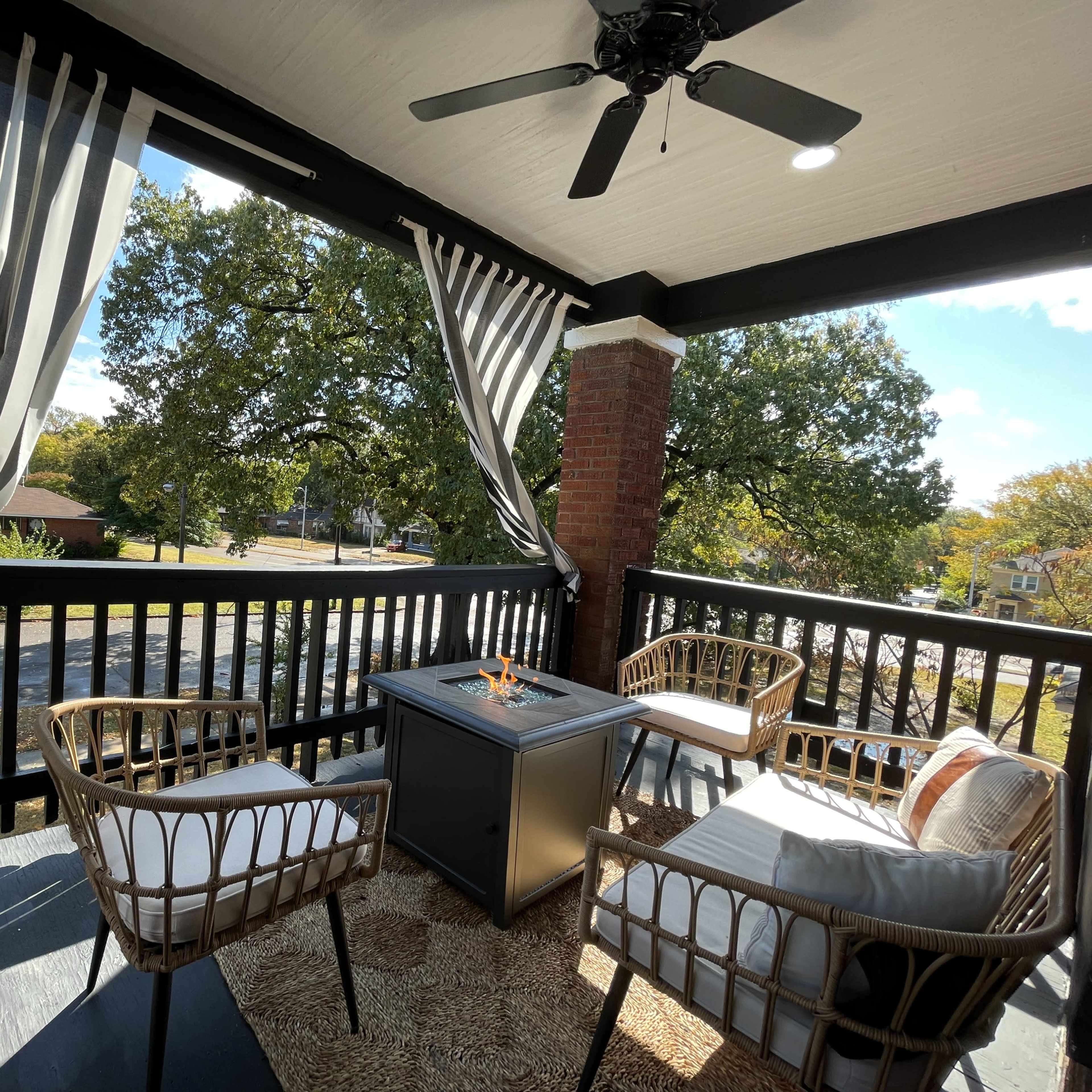 A cozy balcony features four wicker chairs surrounding a fire pit, with striped curtains fluttering in the breeze against a backdrop of trees and a street view.