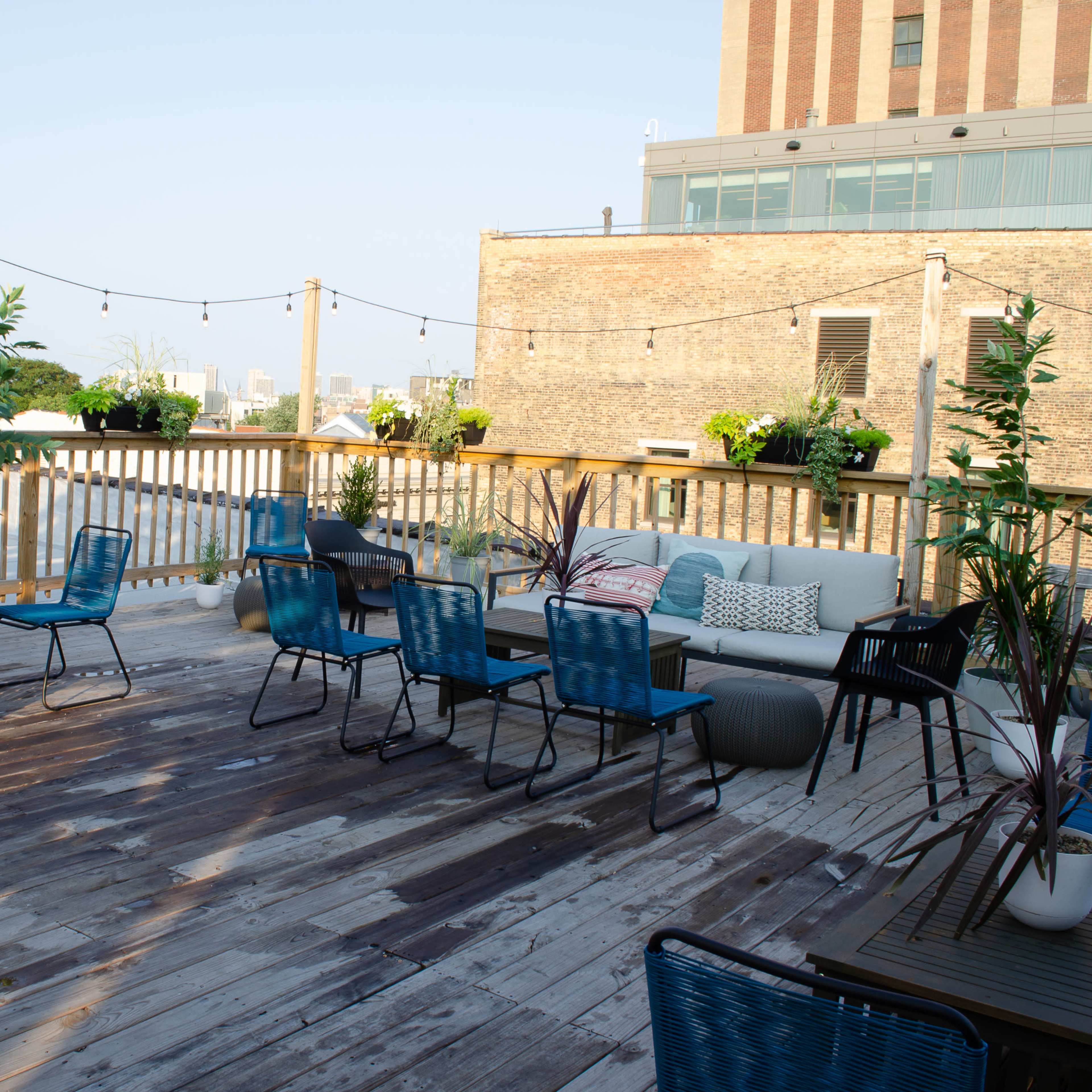 A rooftop deck with wooden flooring, various seating arrangements, potted plants, and string lights above.