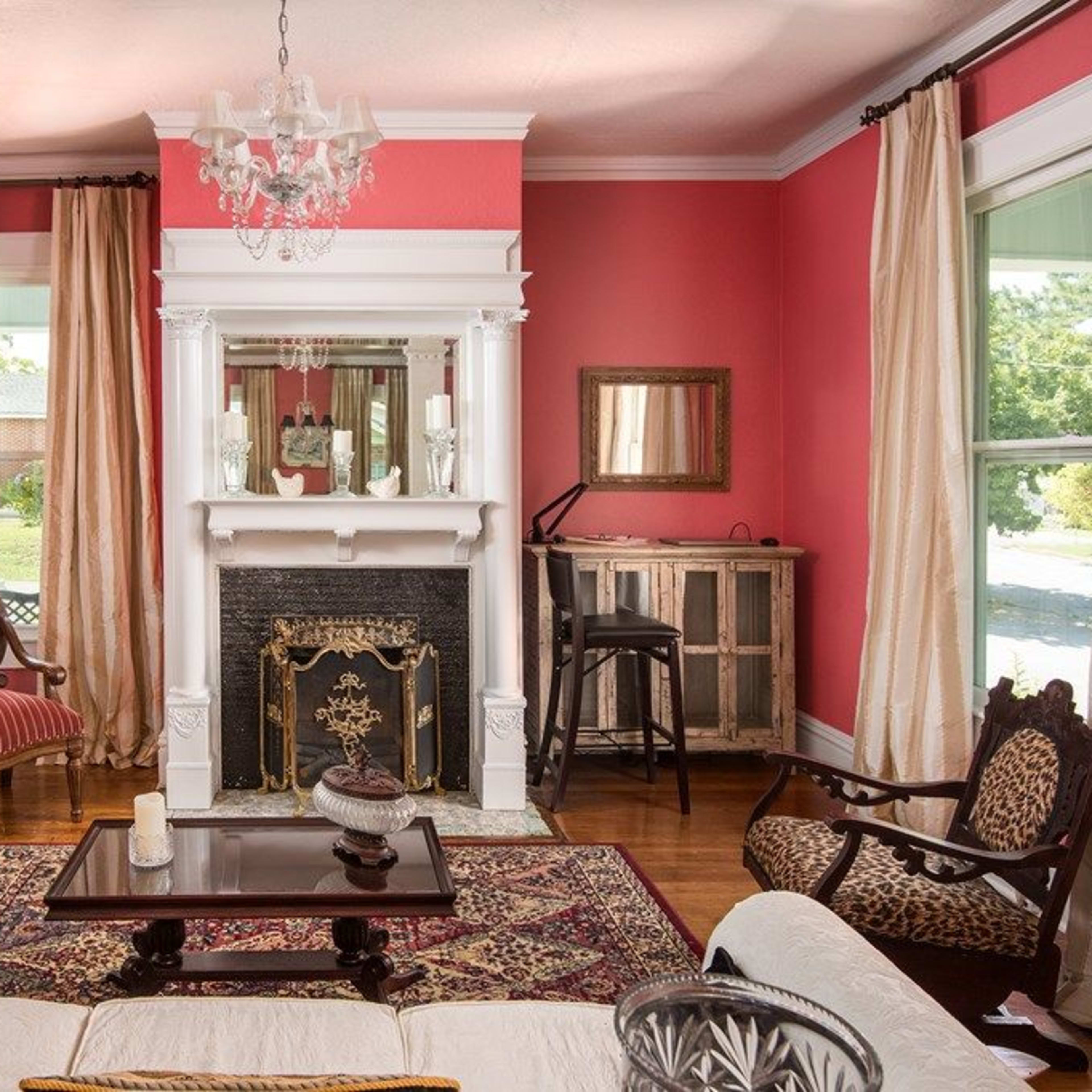 The room features a pink wall, a fireplace with a mirror above it, two armchairs, and a small table across from a sofa, all illuminated by natural light.
