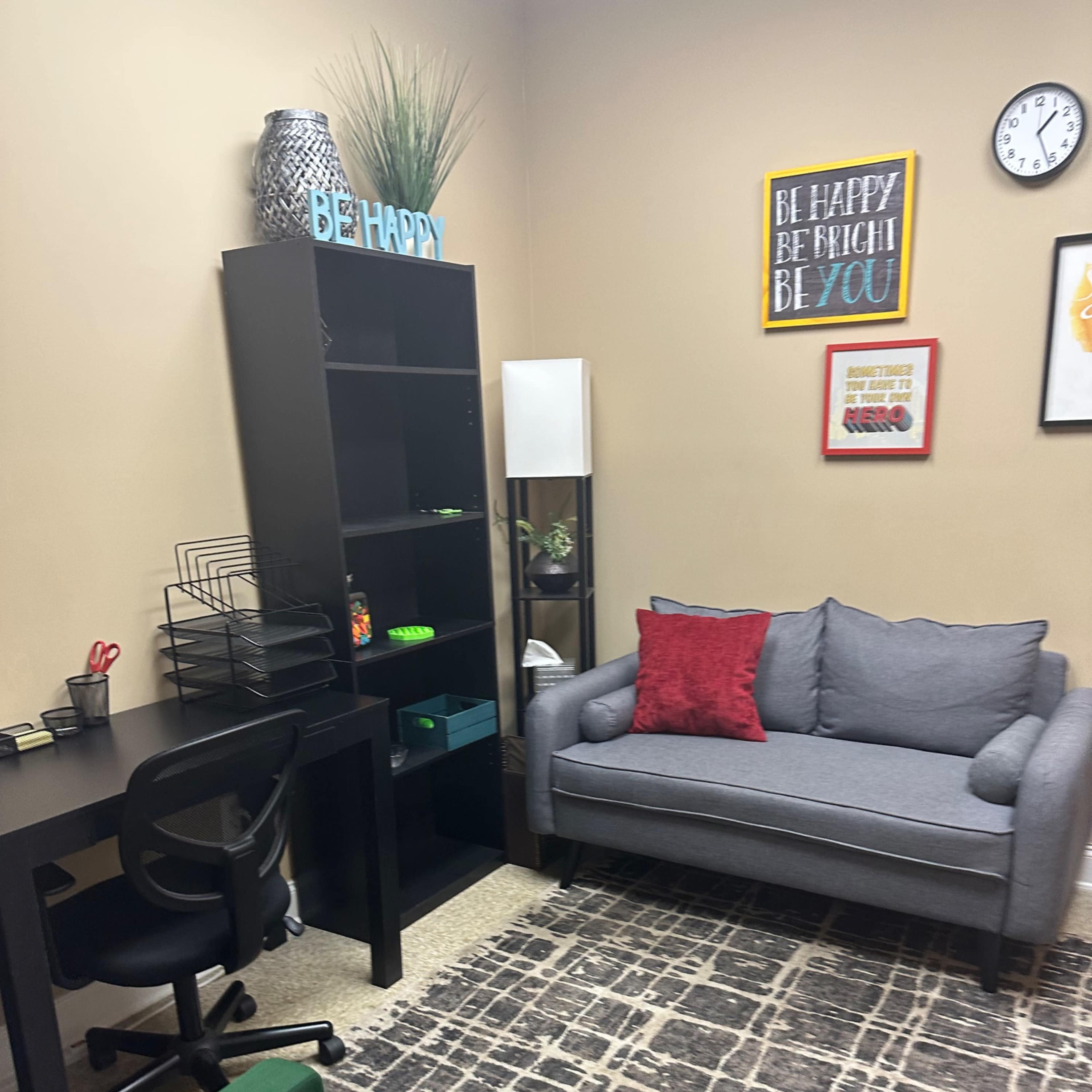 A small office space featuring a gray couch, a green chair, a black desk with a rolling chair, a black bookshelf, and decorations on the walls.