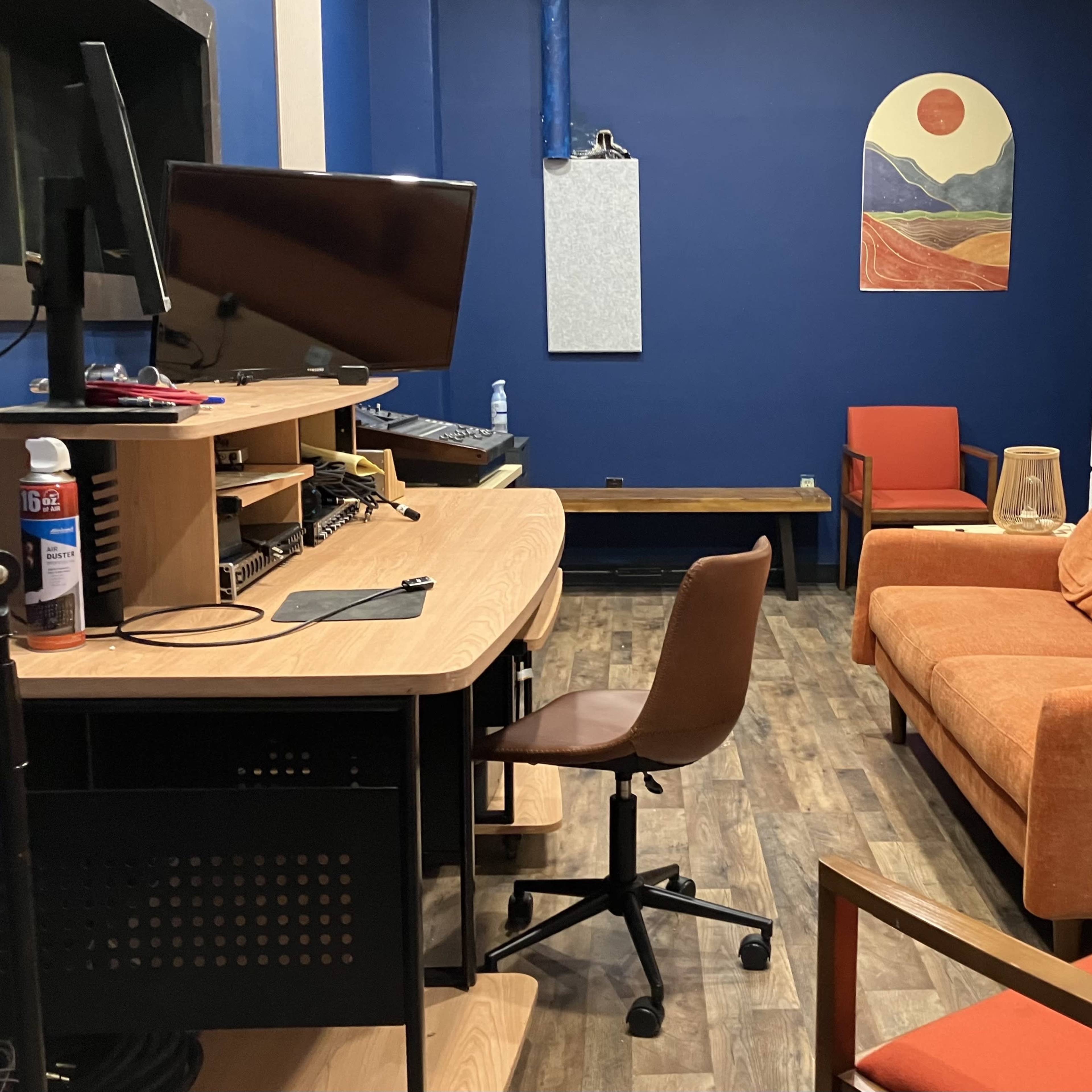 A cozy studio room featuring a wooden desk with a computer, an orange sofa, and artistic wall decor, all set against a blue backdrop.