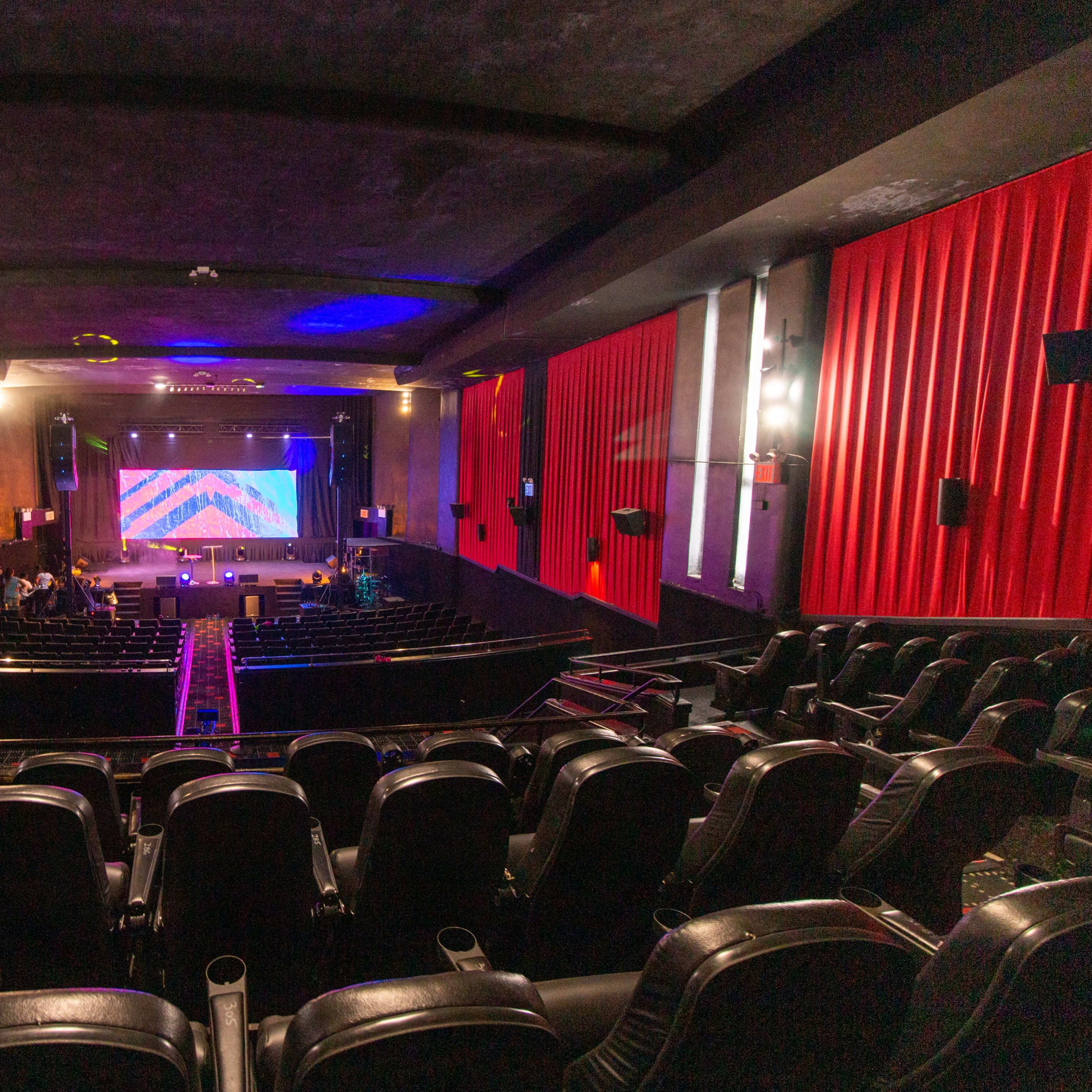 A view from the back of a theater featuring rows of empty black seats facing a stage with a colorful backdrop and red curtains.