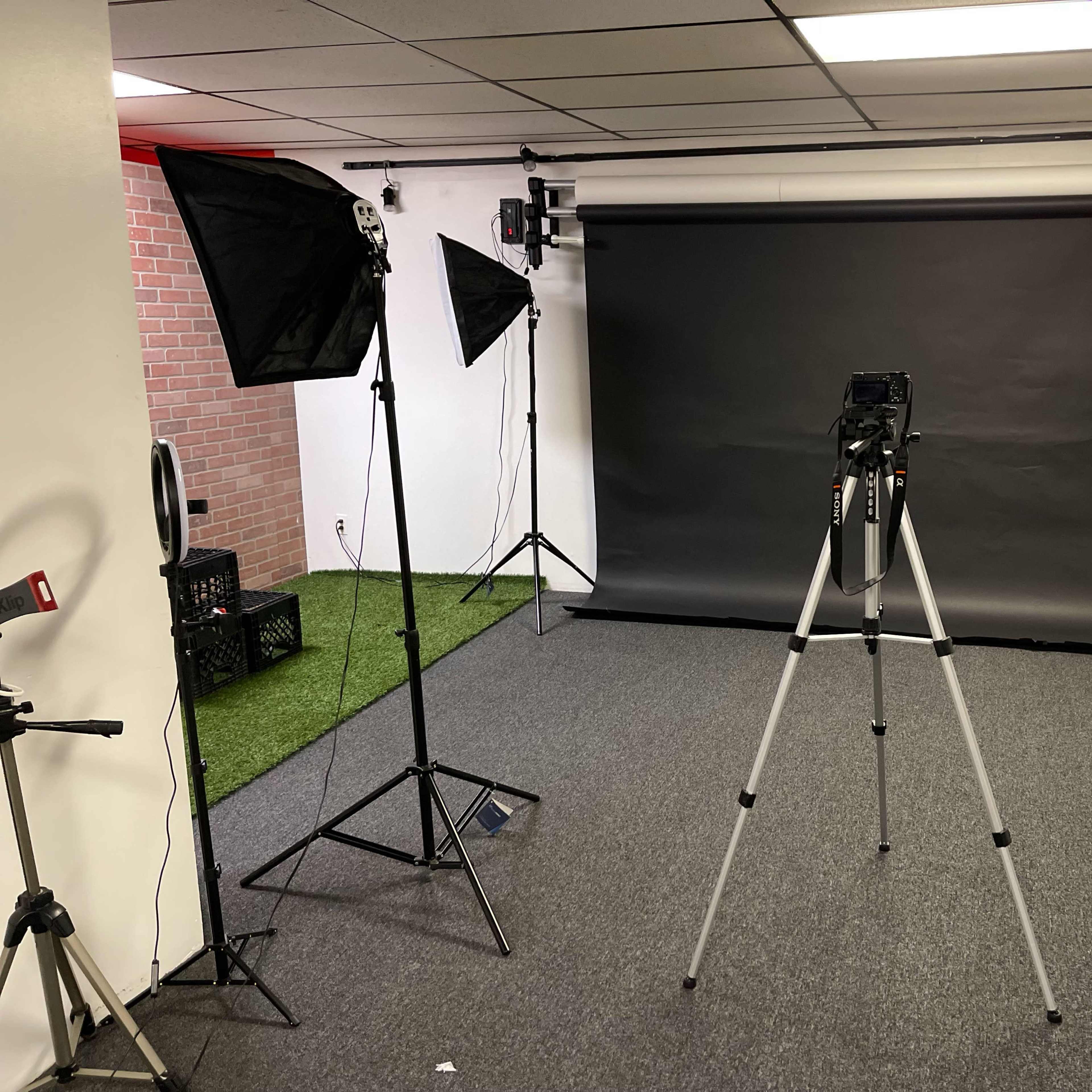A photography studio setup with a black backdrop, a camera on a tripod, and two softbox lights on either side.