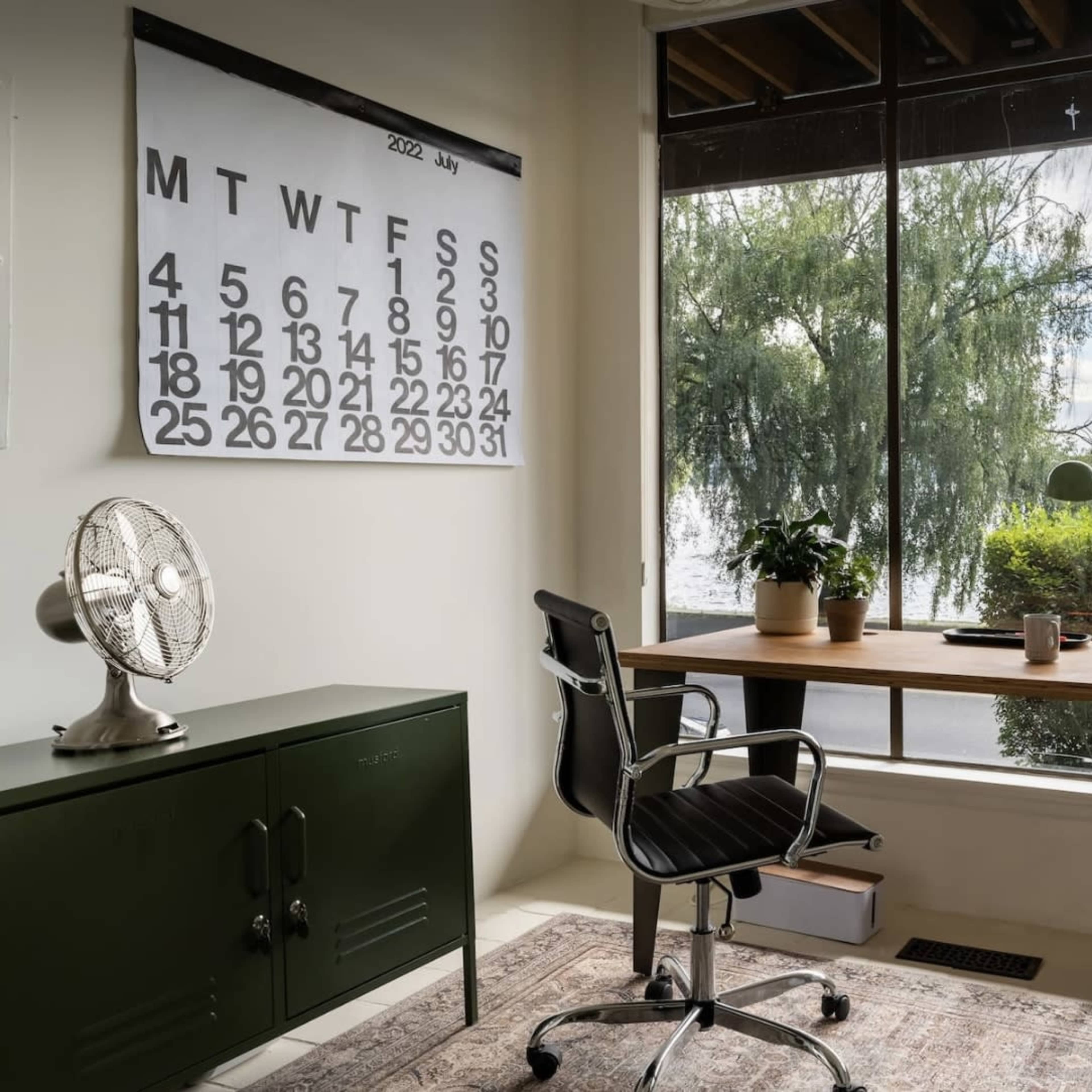 A workspace with a wooden desk, a chair, a calendar mounted on the wall, and large windows overlooking a body of water and greenery.