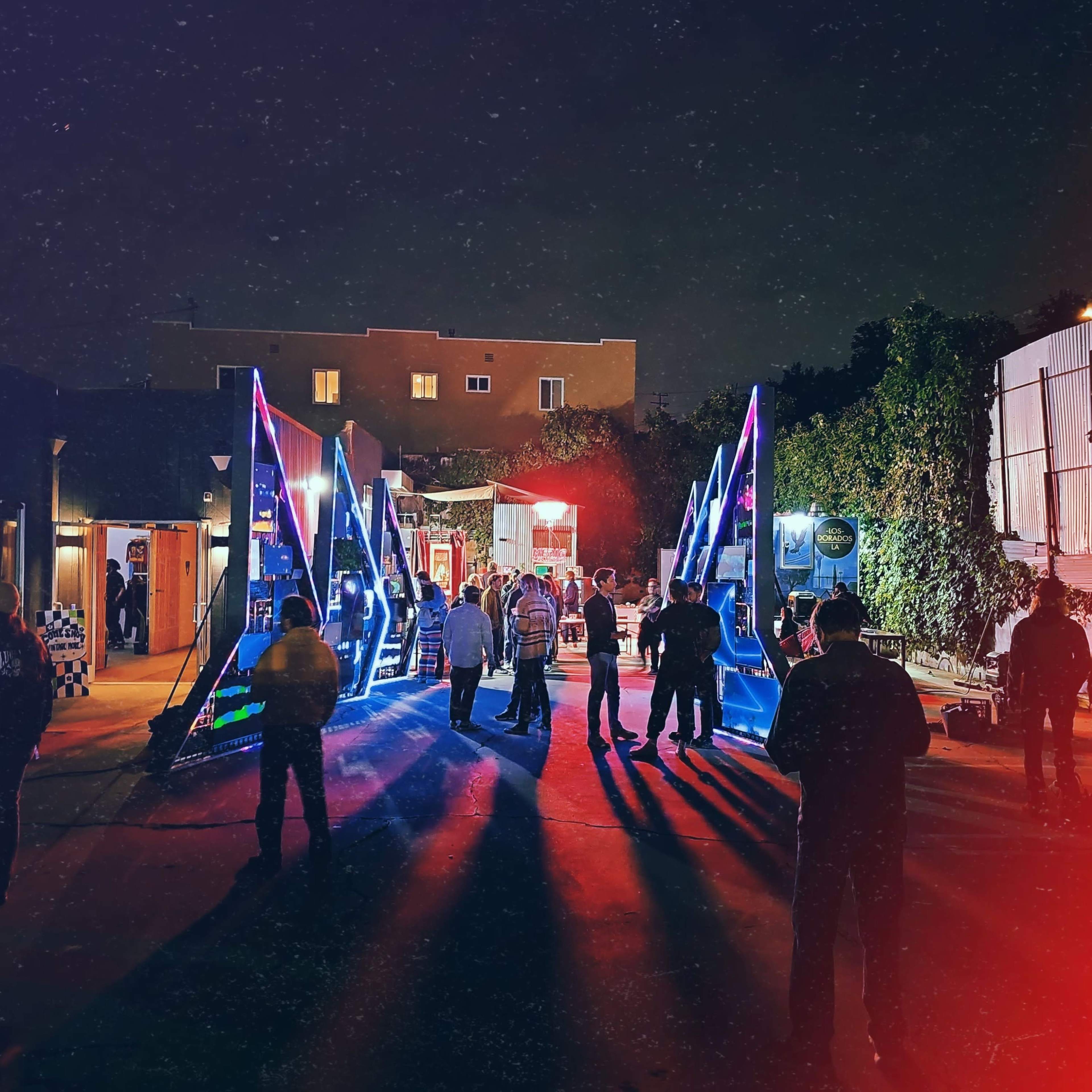 A group of people gathers in a brightly lit area with colorful lighting at night, near large, triangular structures.