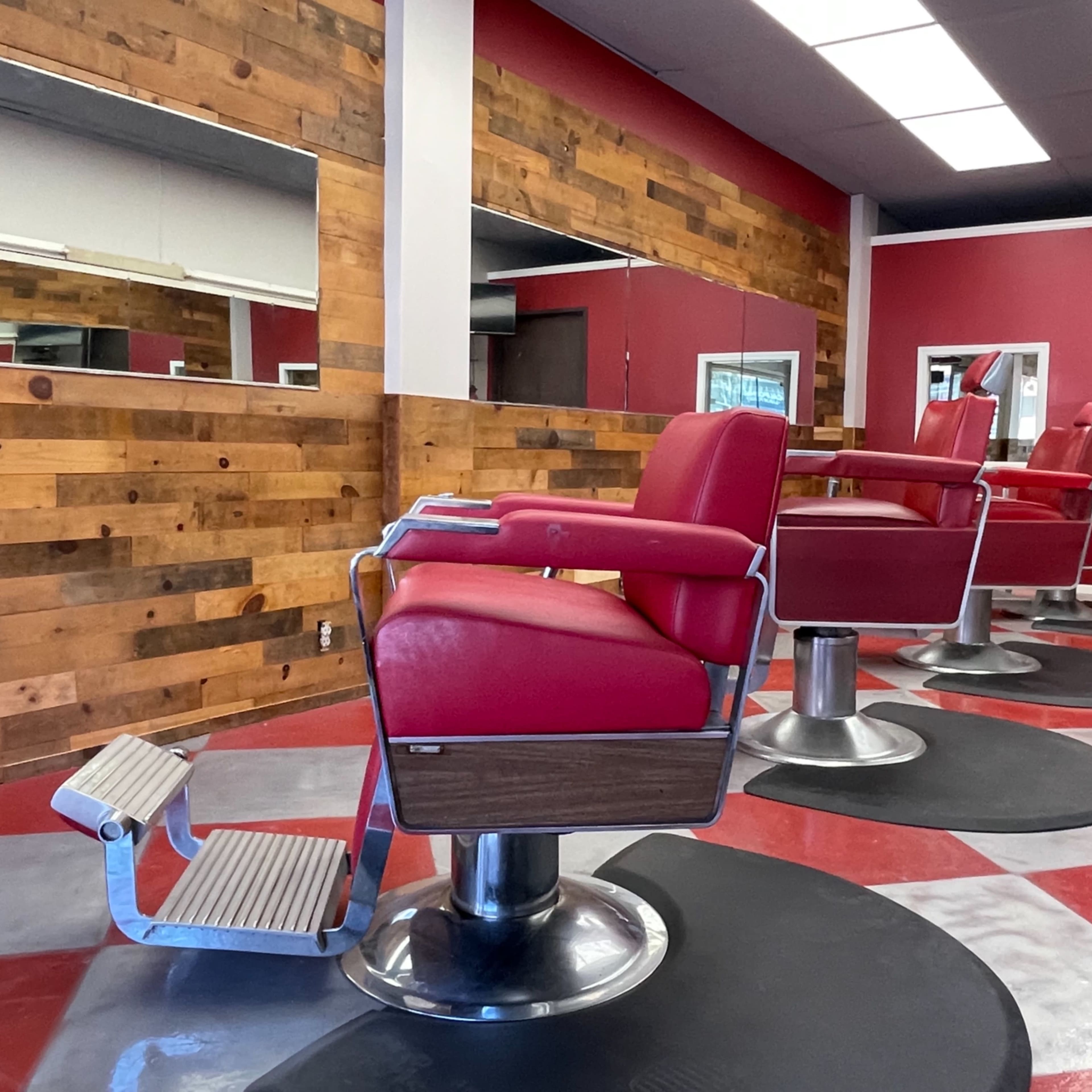 A modern barber shop interior with red and gray flooring, wooden accents on the walls, and several red barber chairs positioned in front of mirrors.