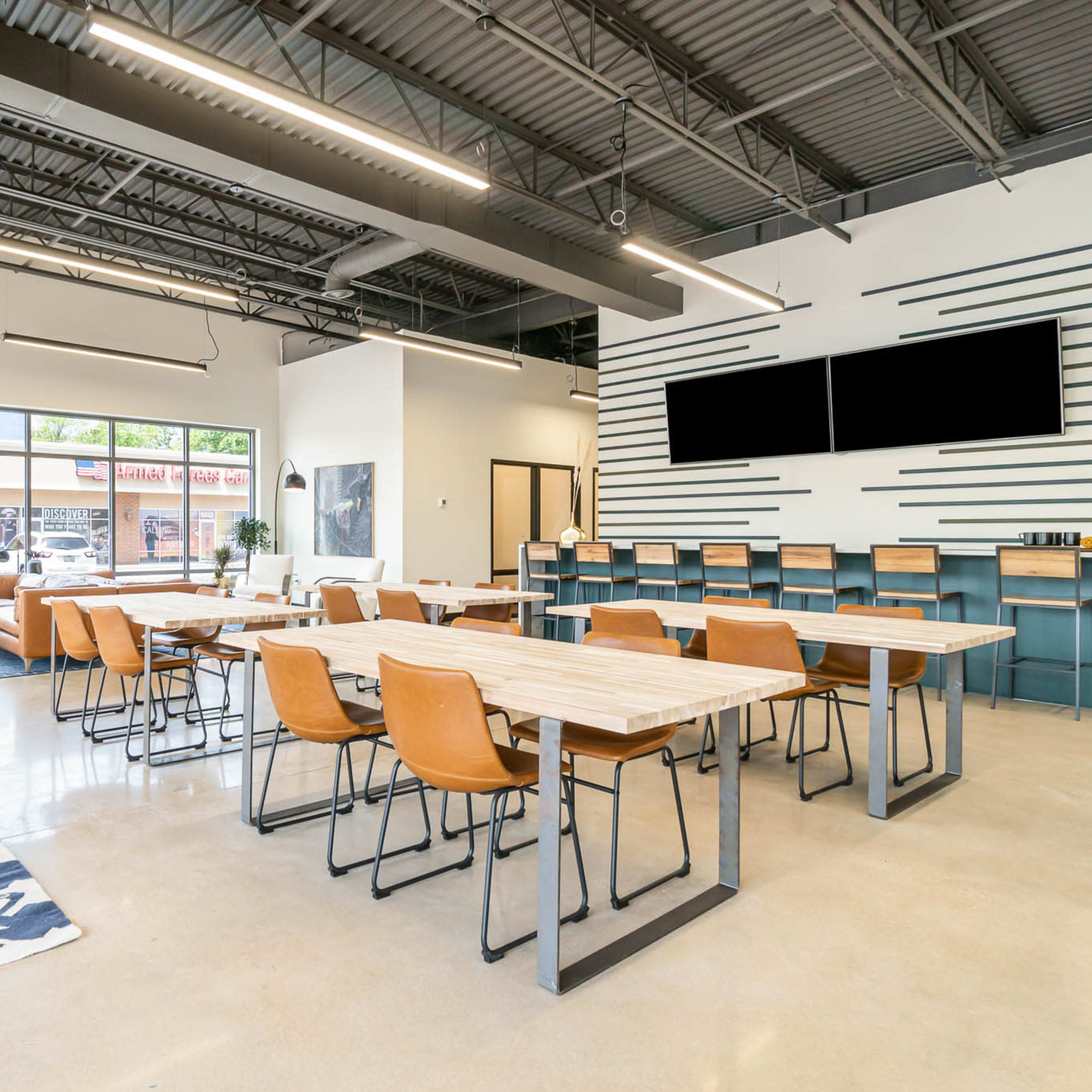 A spacious, modern work area features multiple tables and chairs, large windows letting in natural light, and a seating area with a plant.