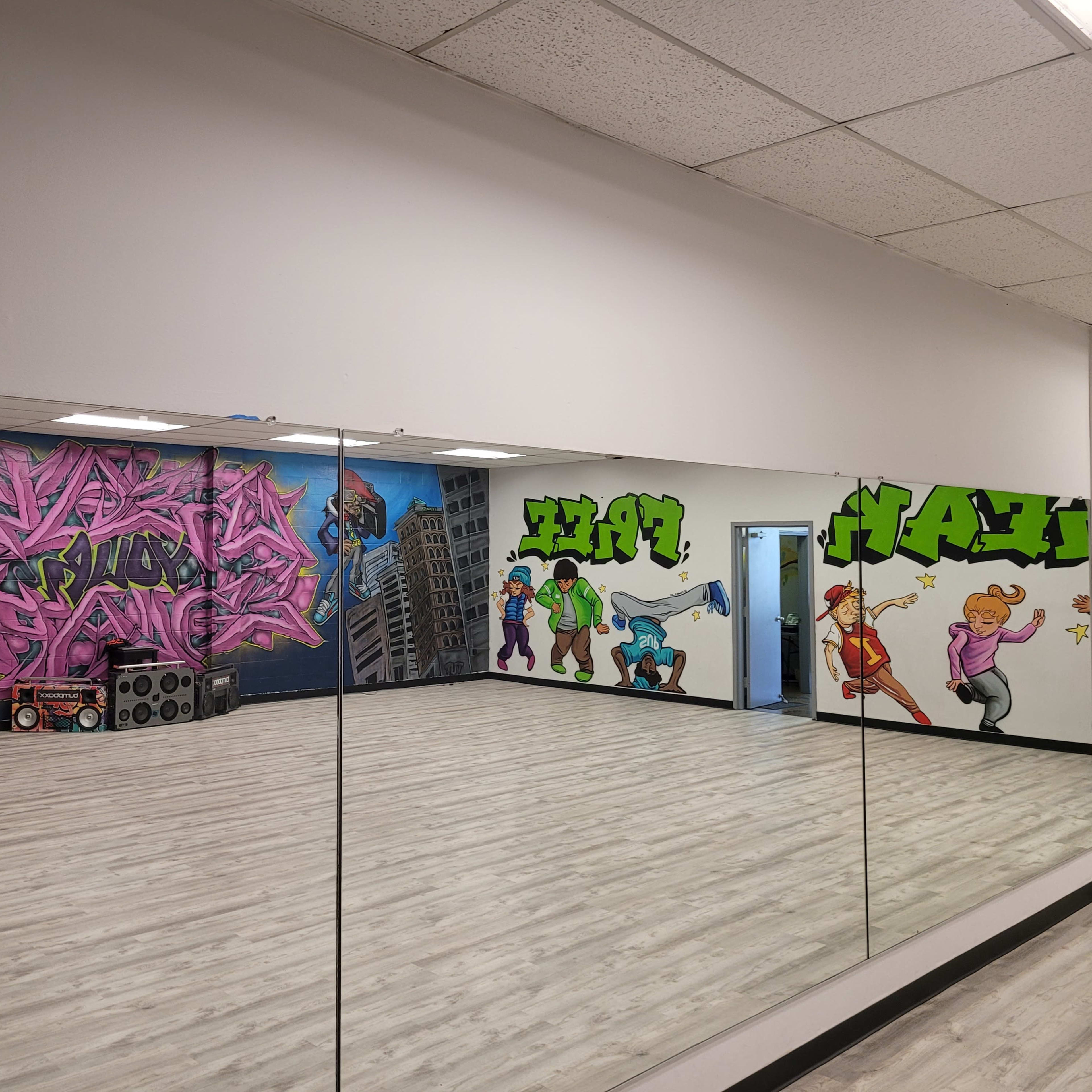A dance studio with a large mirror, featuring colorful graffiti murals on the walls and wooden flooring.