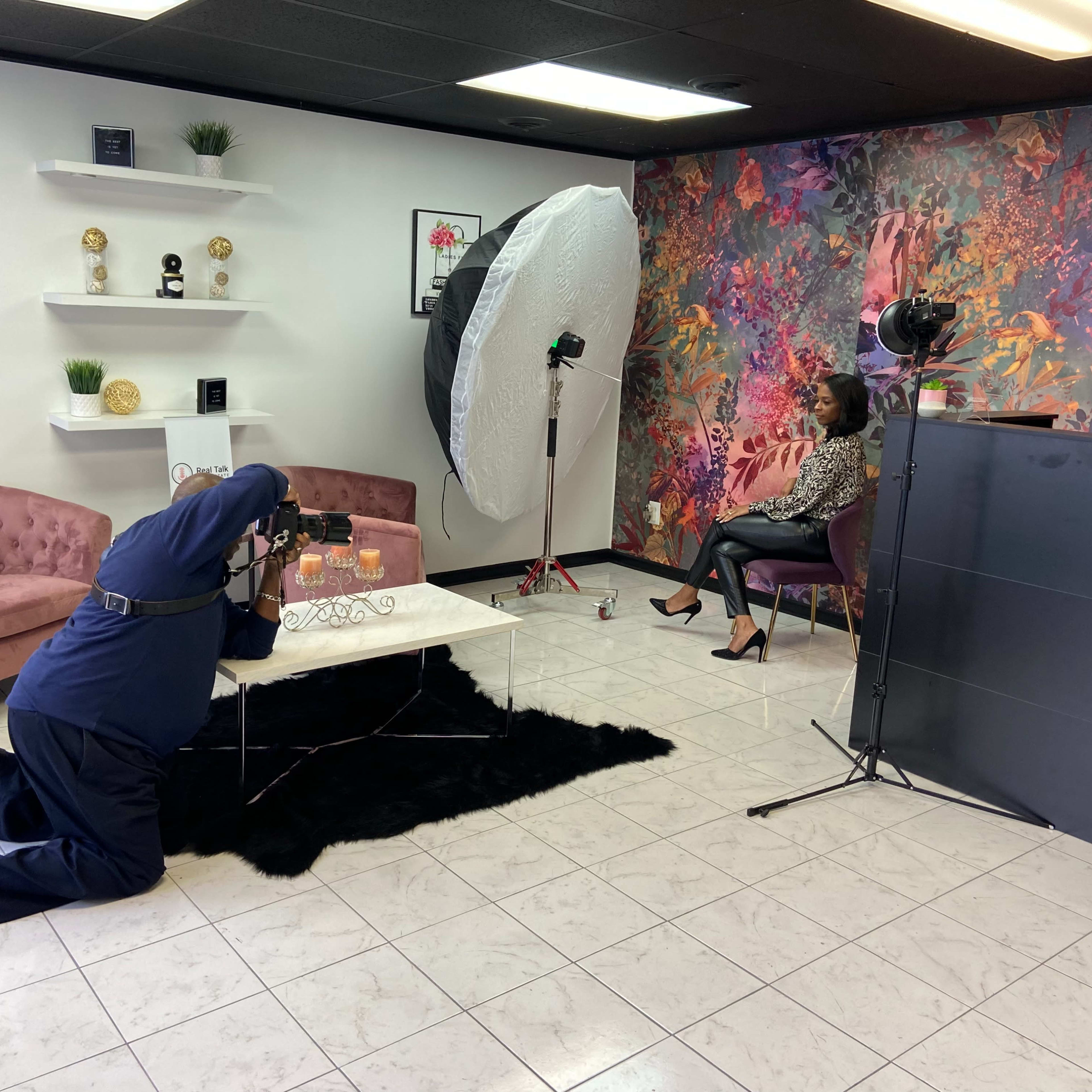 A photographer is kneeling on the floor while capturing a portrait of a seated model in a studio decorated with flowers and modern furnishings.