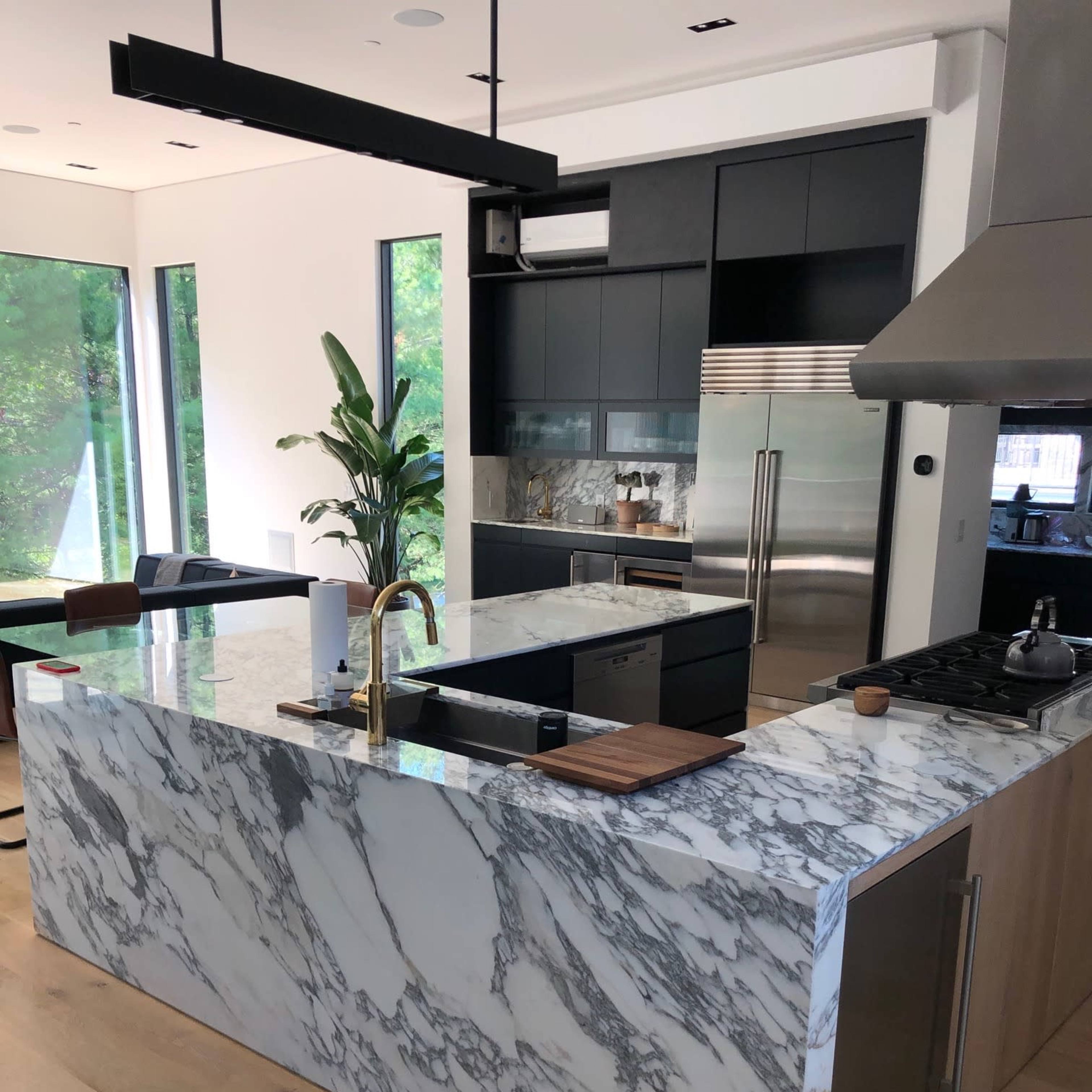 A modern kitchen with a large marble island, a stainless steel stove, and floor-to-ceiling windows featuring a view of greenery outside.