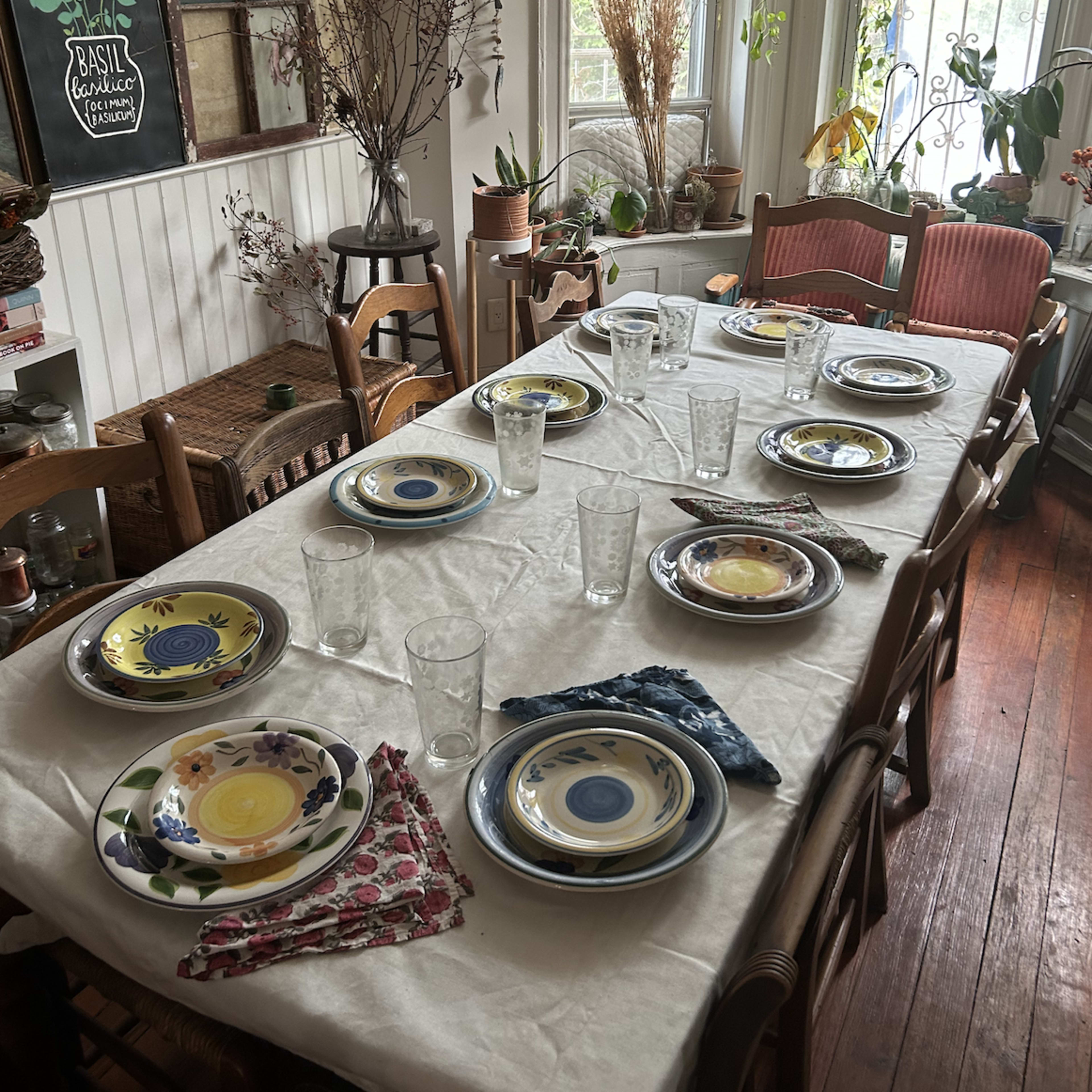 A long dining table is set with various plates and glasses in a well-lit room filled with plants and decorative items.