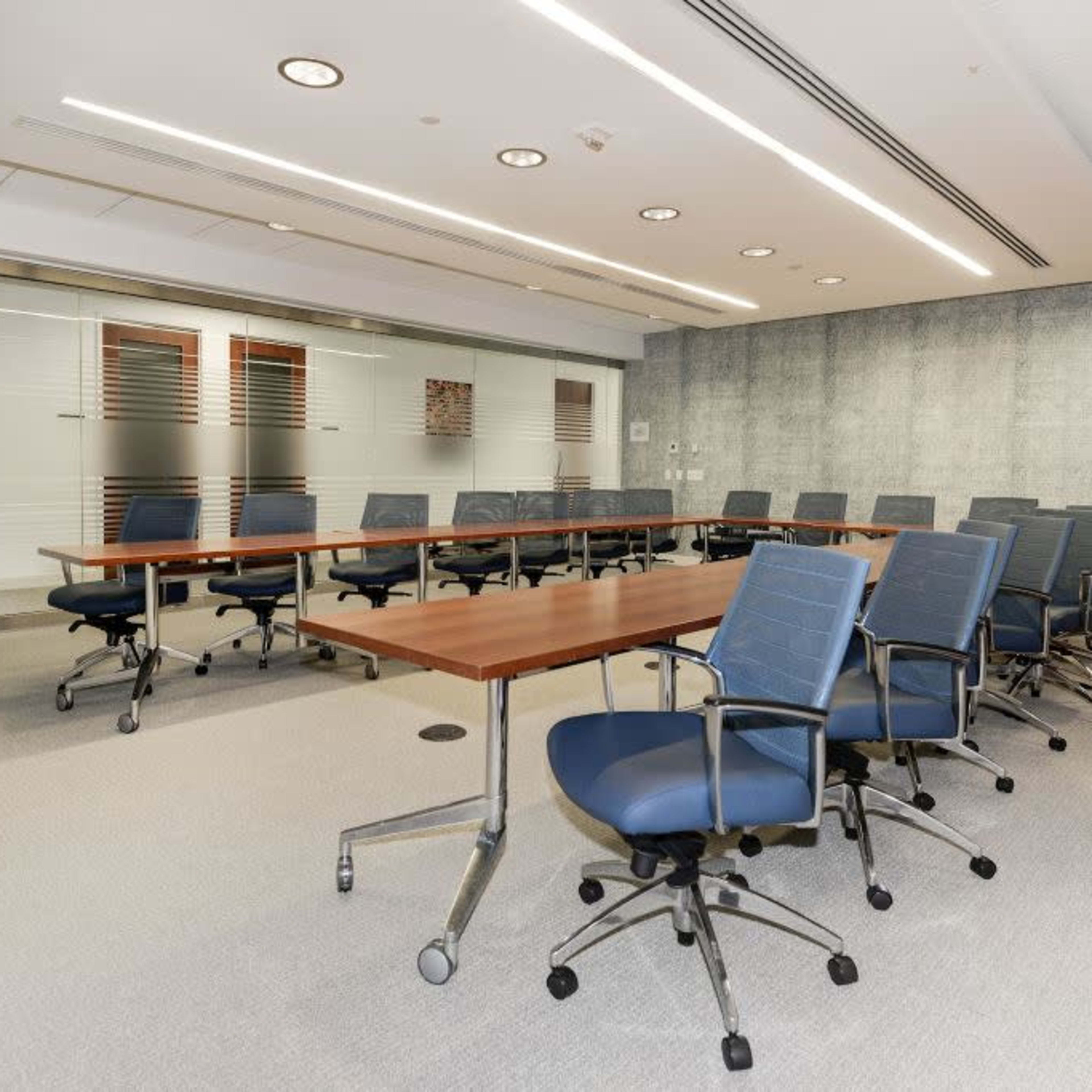 A modern conference room features a long wooden table surrounded by blue ergonomic chairs, with glass panels and a textured wall.