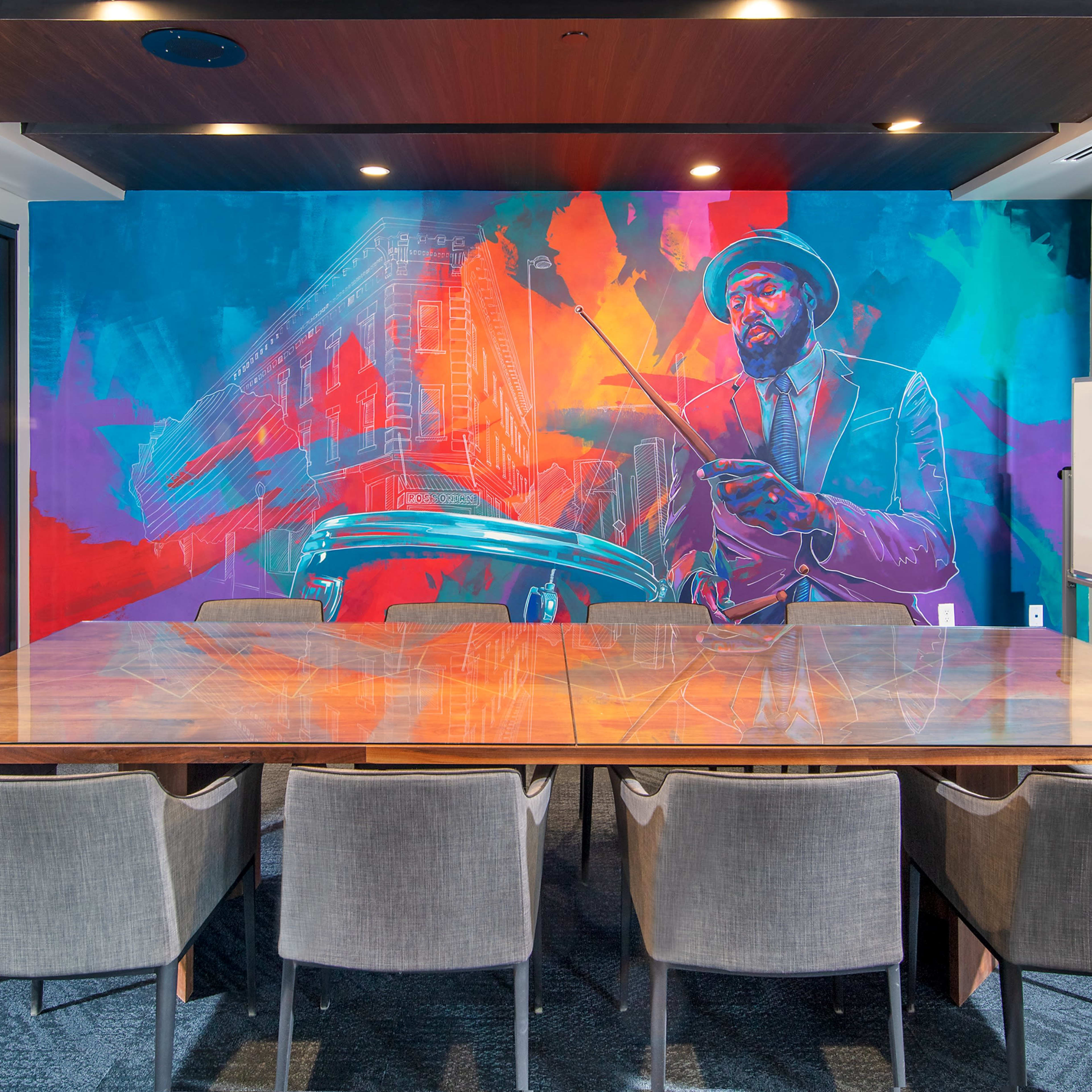 A modern conference room features a large wooden table surrounded by gray chairs, with a vibrant mural of a man holding a conductor's baton on the wall.