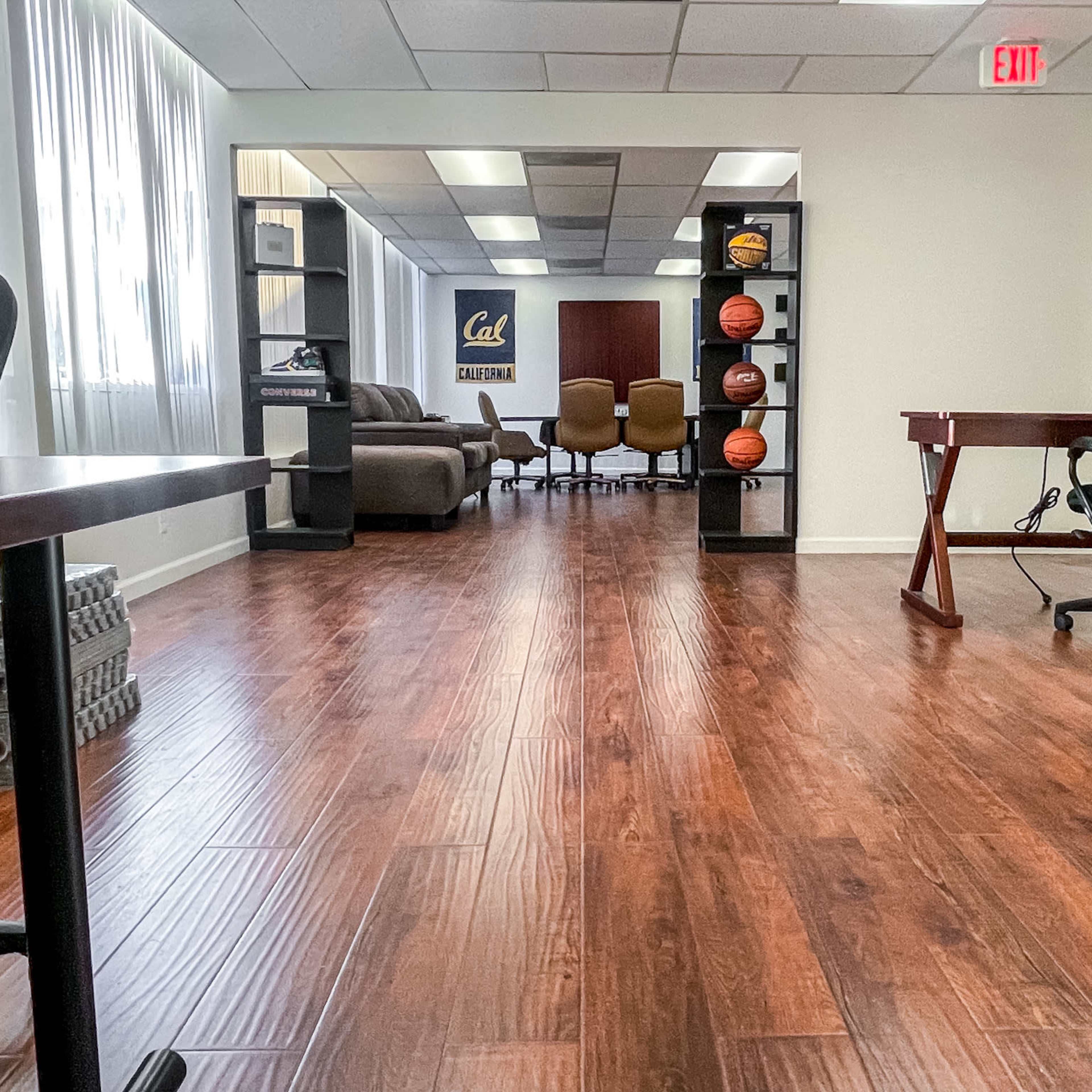 A modern office space with wooden flooring, featuring desks on either side and a meeting area with chairs and basketballs displayed on shelves in the background.