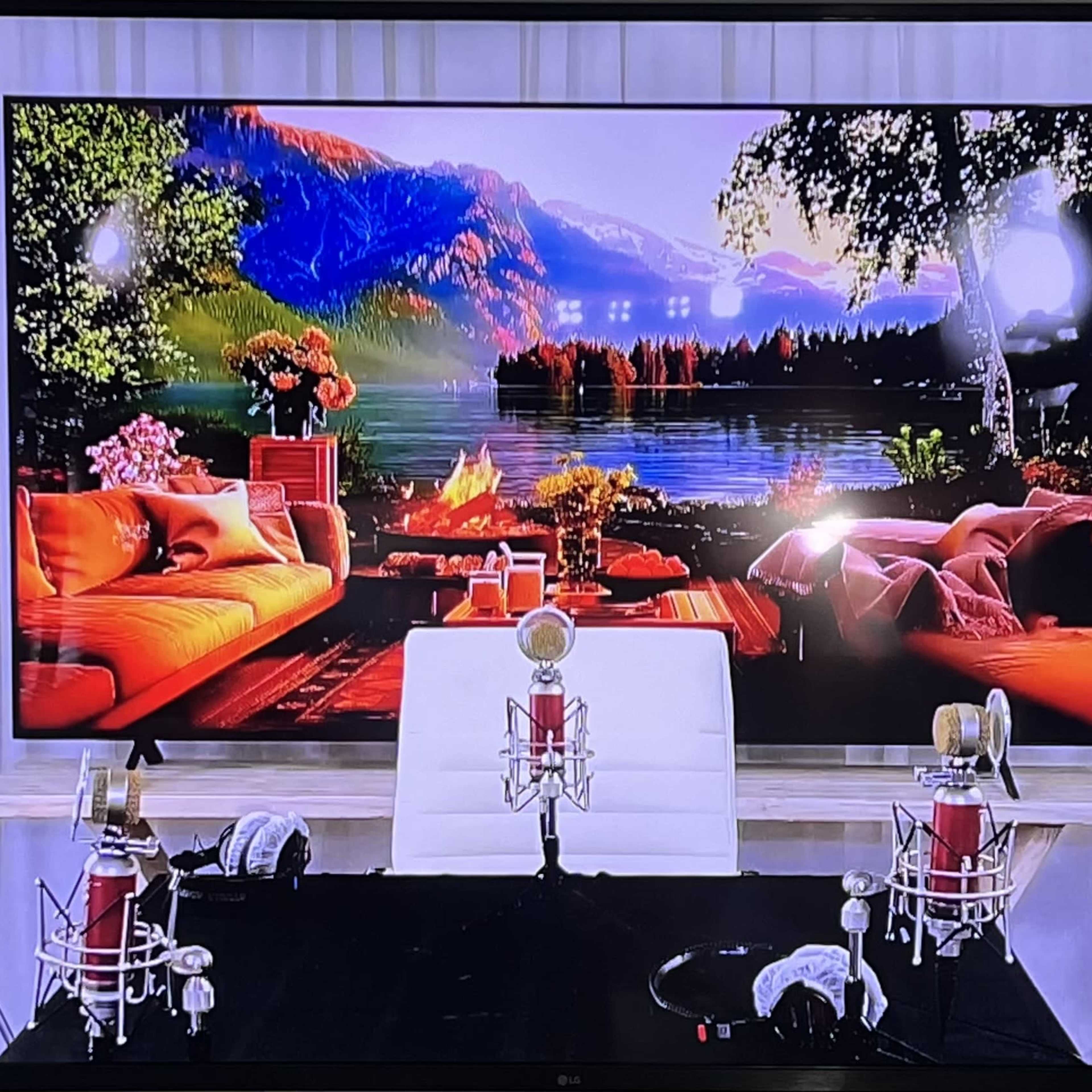 A large screen displaying a scenic landscape, flanked by two cameras and set up with microphones on a stage with a white backdrop.