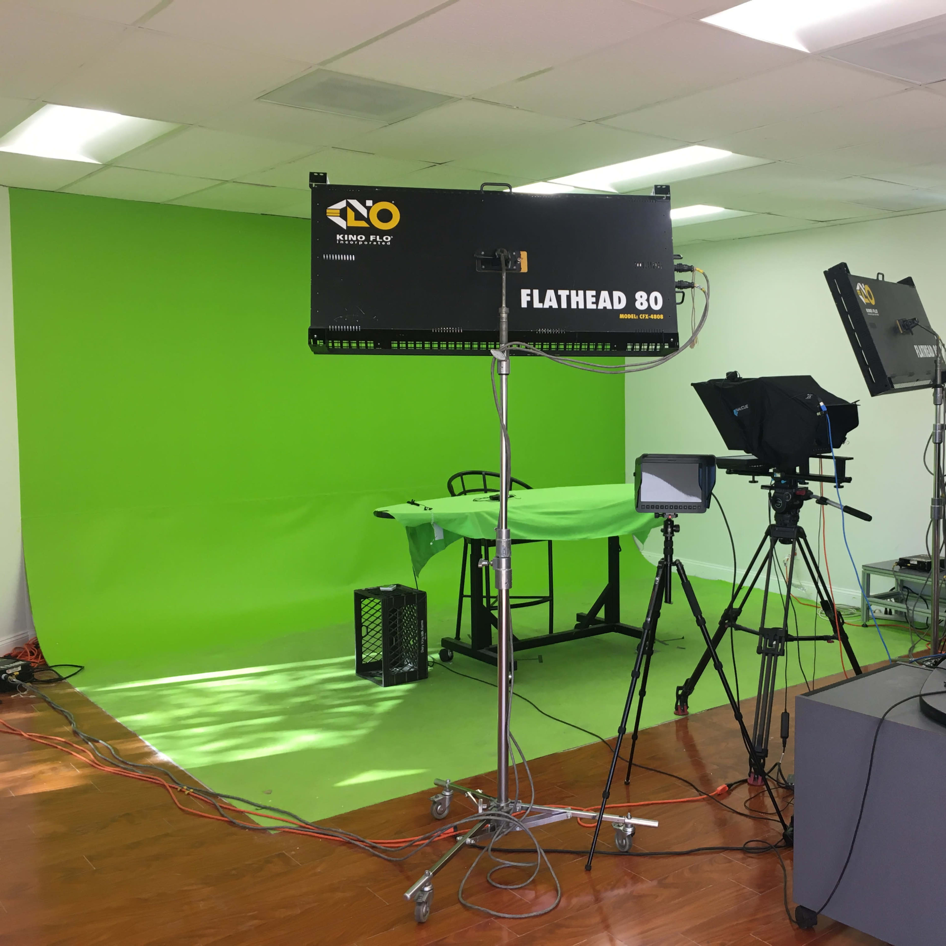 A studio setup featuring a green screen, lighting equipment, and video cameras arranged on a floor with wood finish.