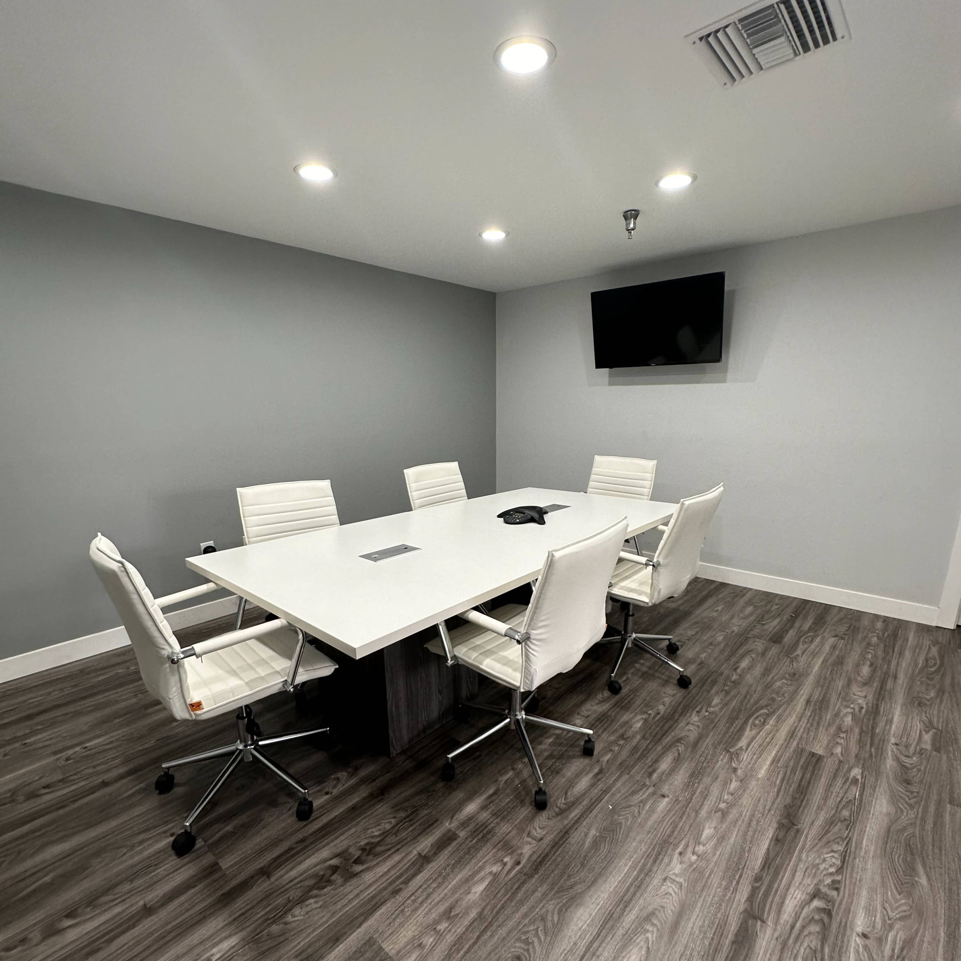 A modern conference room features a large white table surrounded by six ergonomic chairs, with a wall-mounted television and gray walls.
