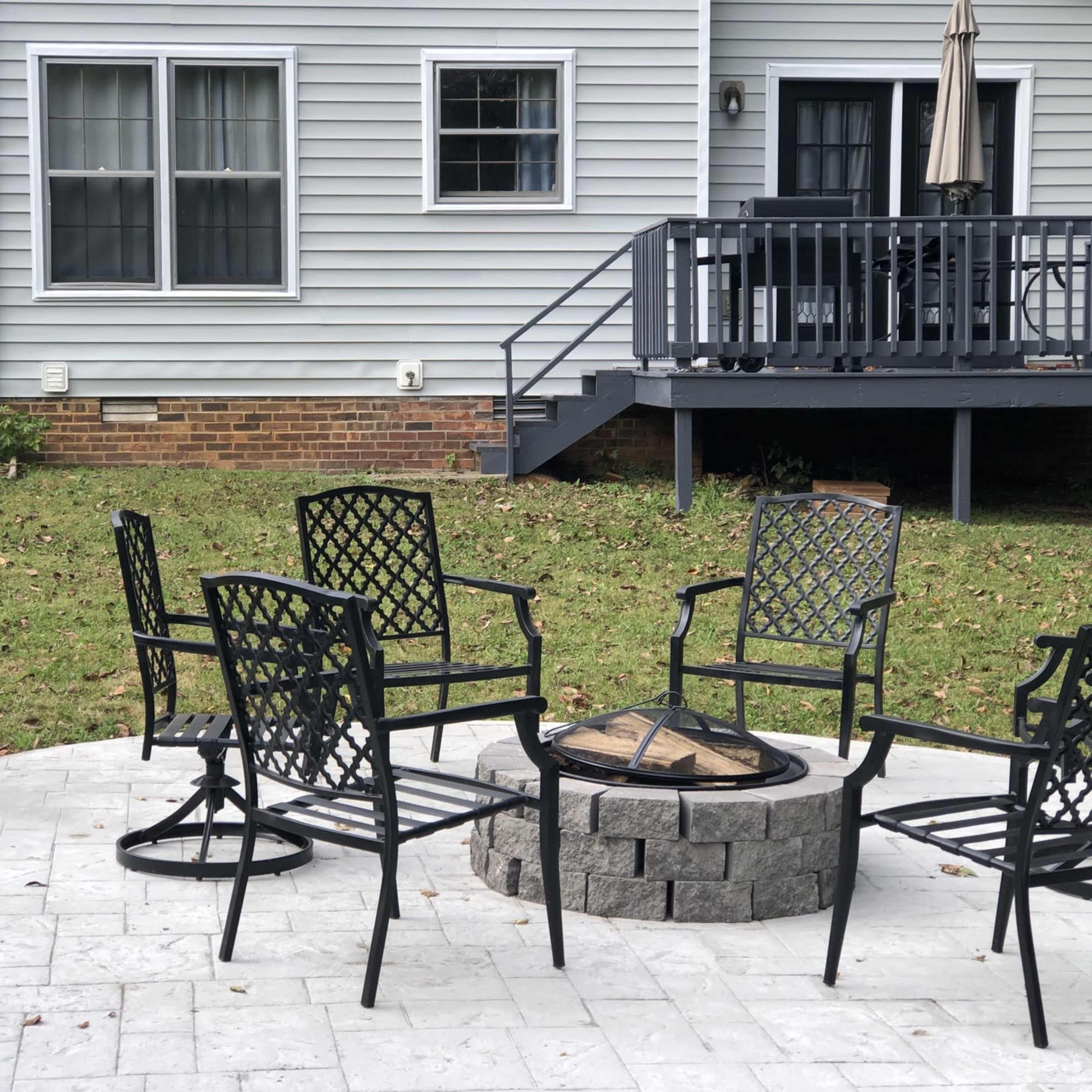 A circular stone fire pit surrounded by five black metal chairs on a paved patio, with a house and a stack of firewood in the background.