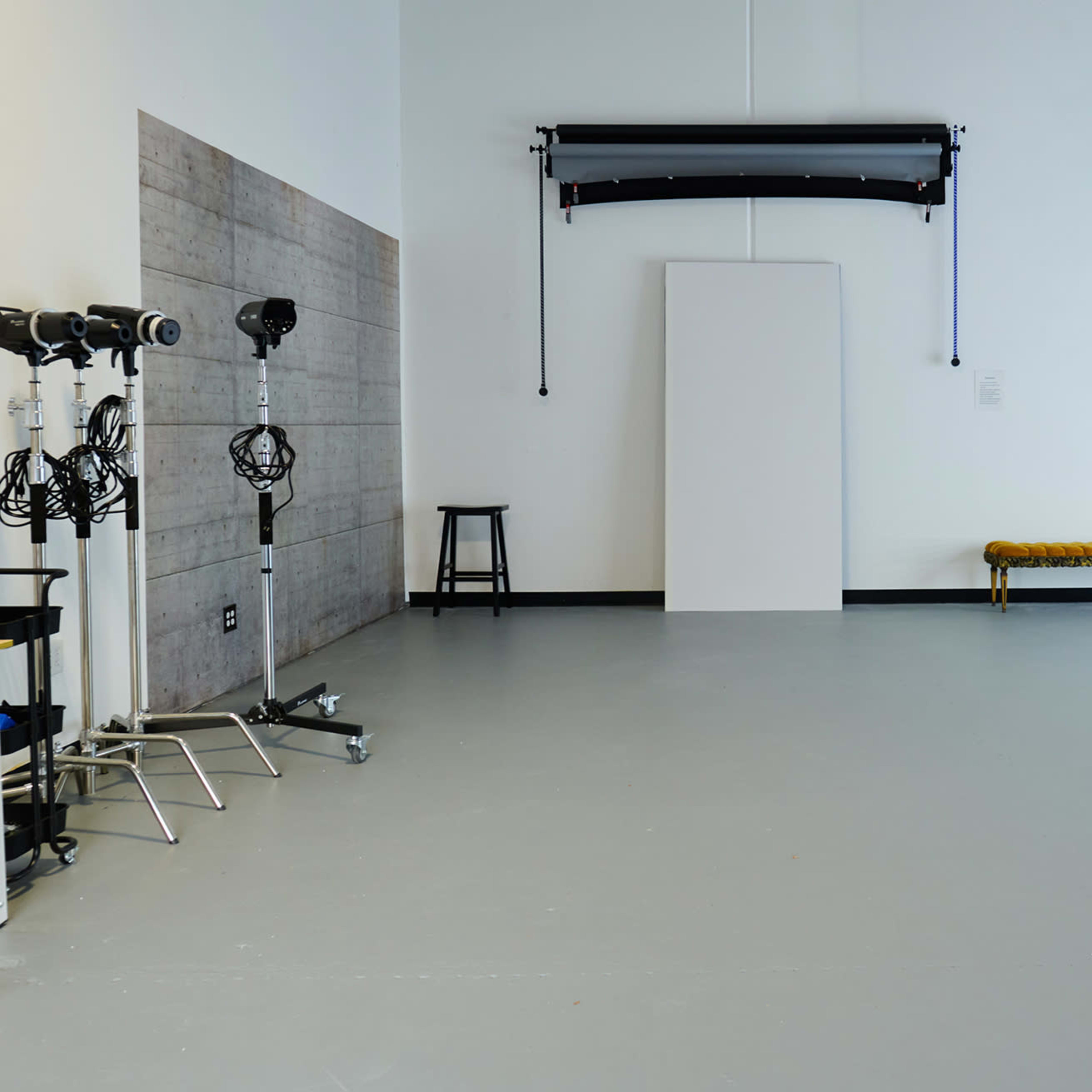 A spacious photography studio with various lighting equipment, a backdrop, and a decorative bench against a plain wall.