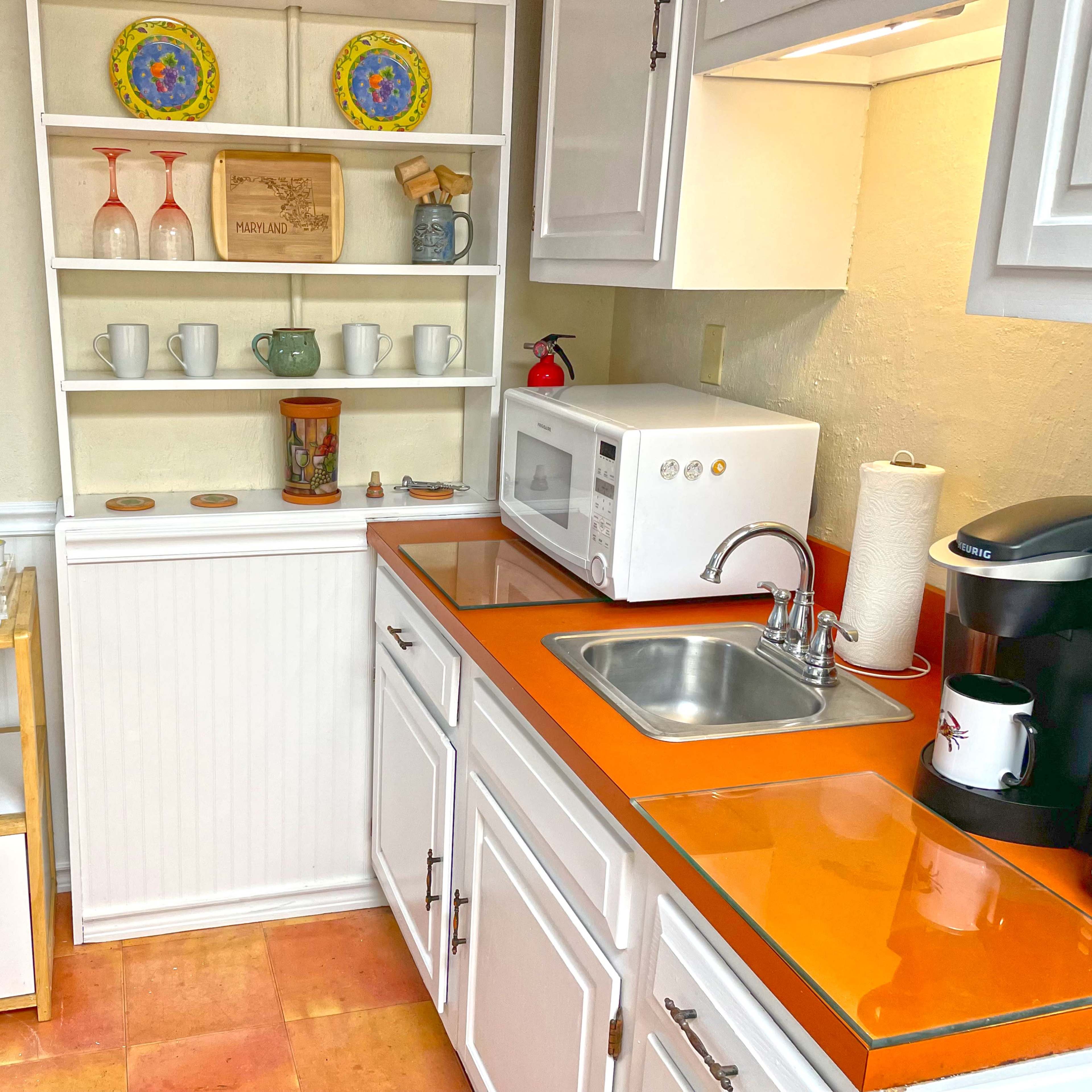 A compact kitchen features white cabinetry, an orange countertop, a microwave, a coffee maker, and open shelving displaying dishes and decorative items.