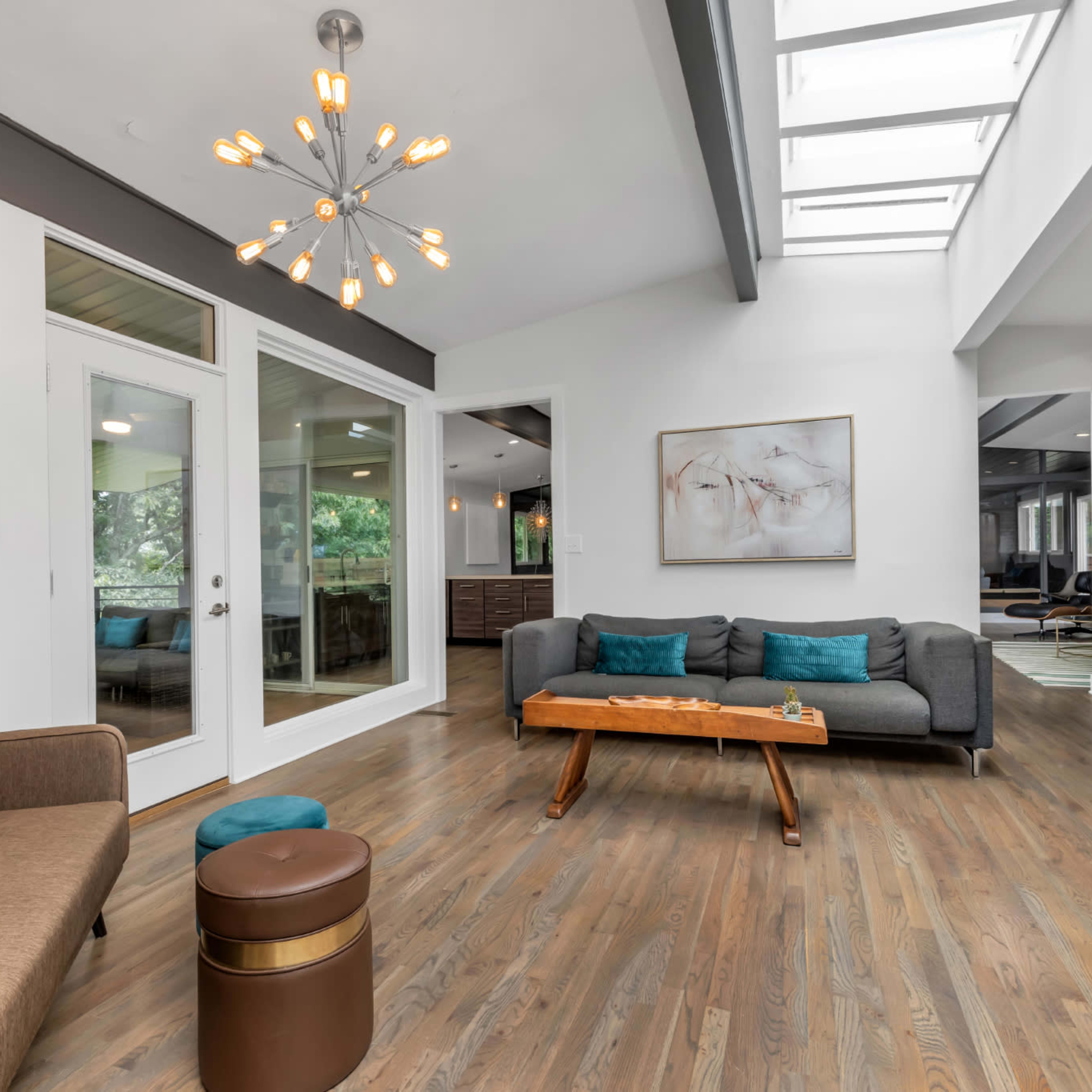 A modern living area featuring a light-filled space with large windows, wooden floors, and contemporary furniture, including a sofa and a coffee table.