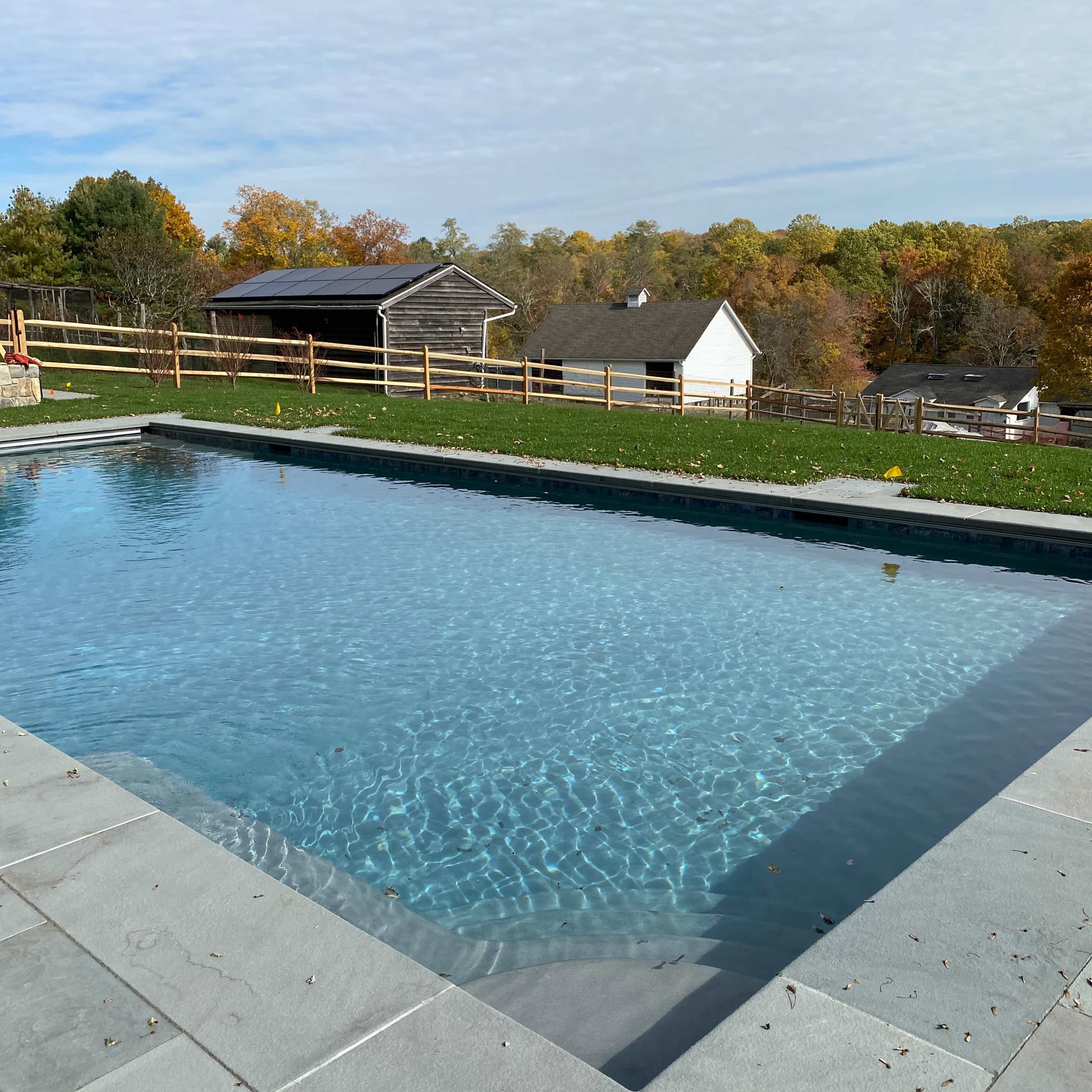 A clear swimming pool next to a grassy area with wooden fences and farm buildings in the background.