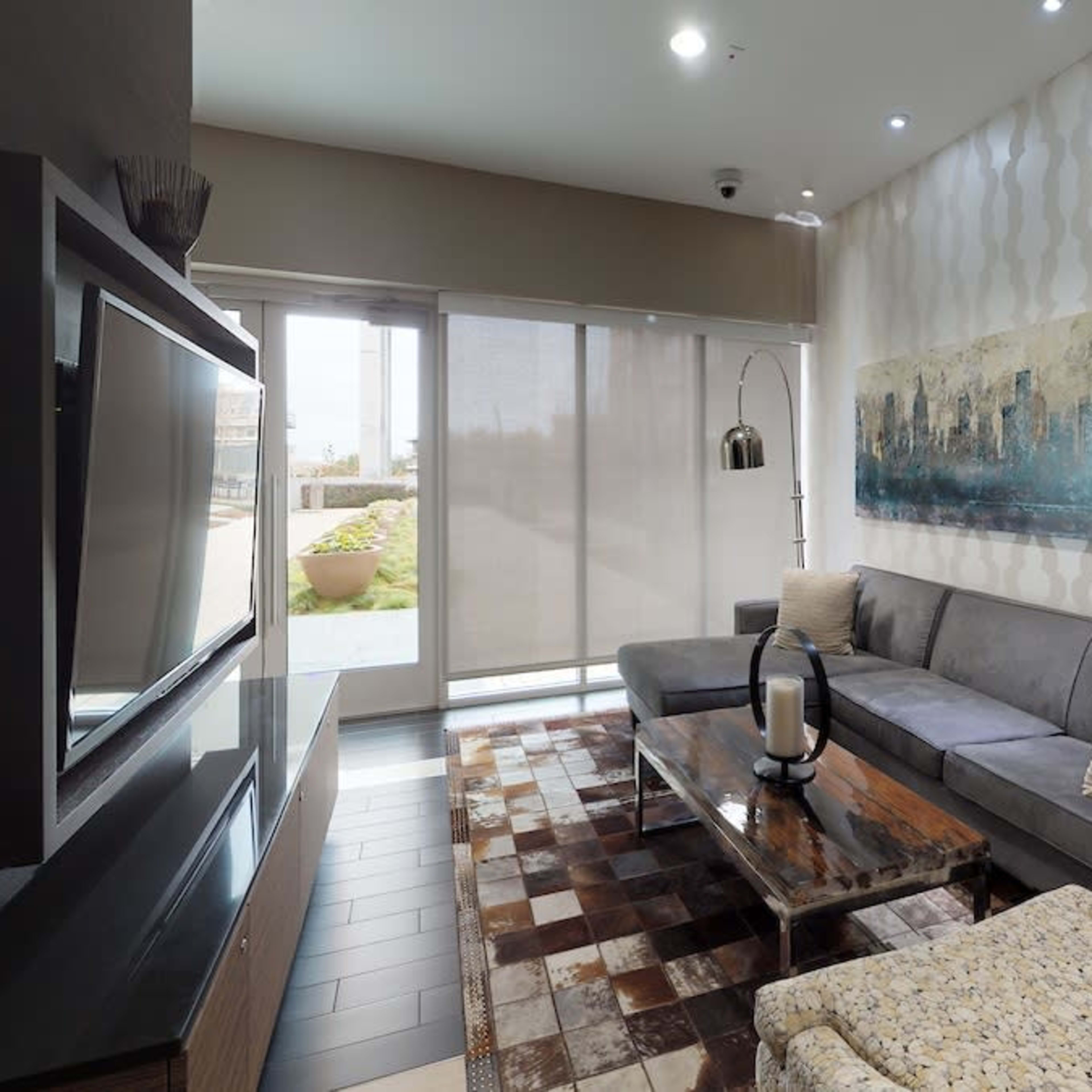A modern living room features a gray sofa, a wooden coffee table, and large windows with blinds, overlooking a landscaped area.