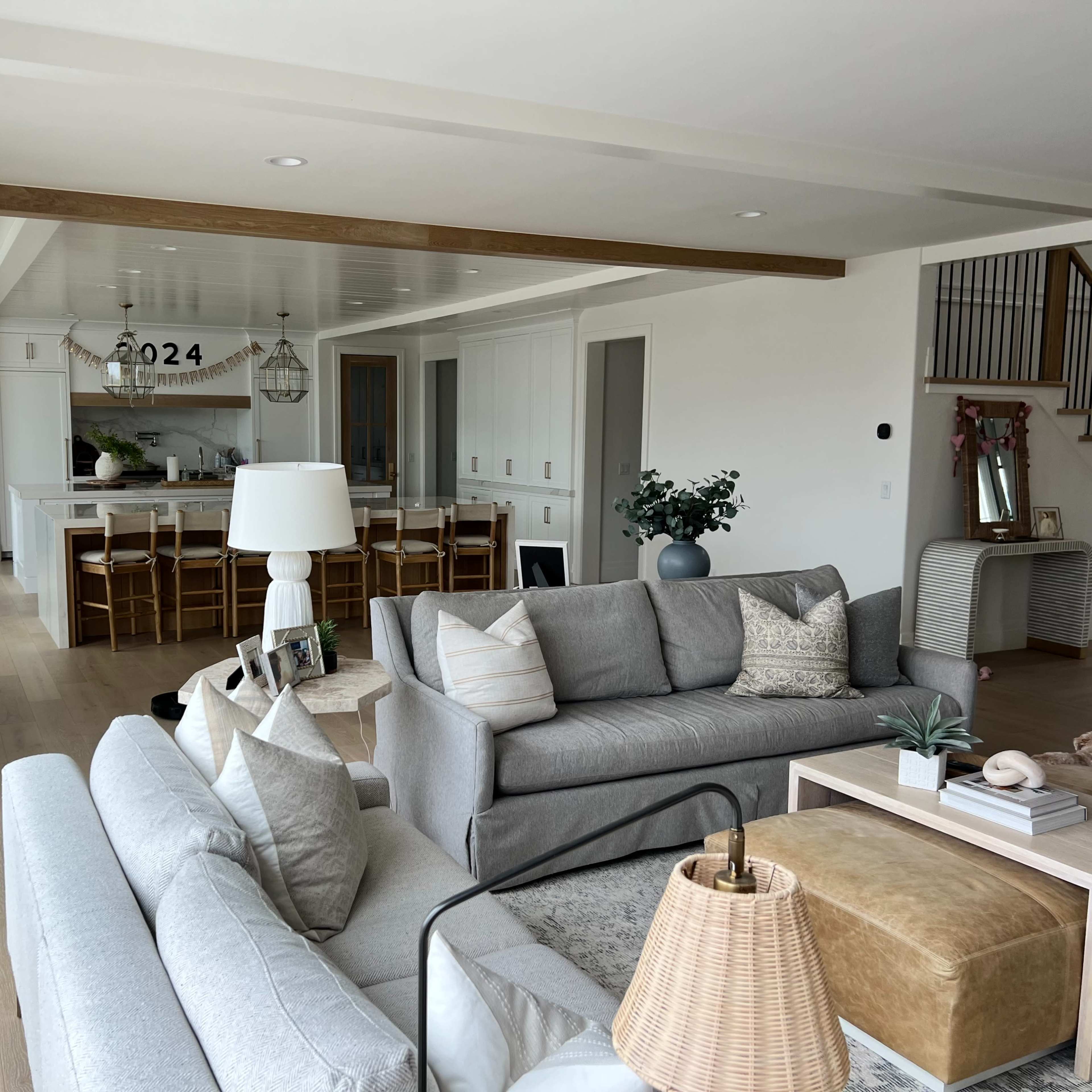 A modern living room with a light-colored sofa, coffee table, and dining area visible in the background, featuring a spacious kitchen and staircase.