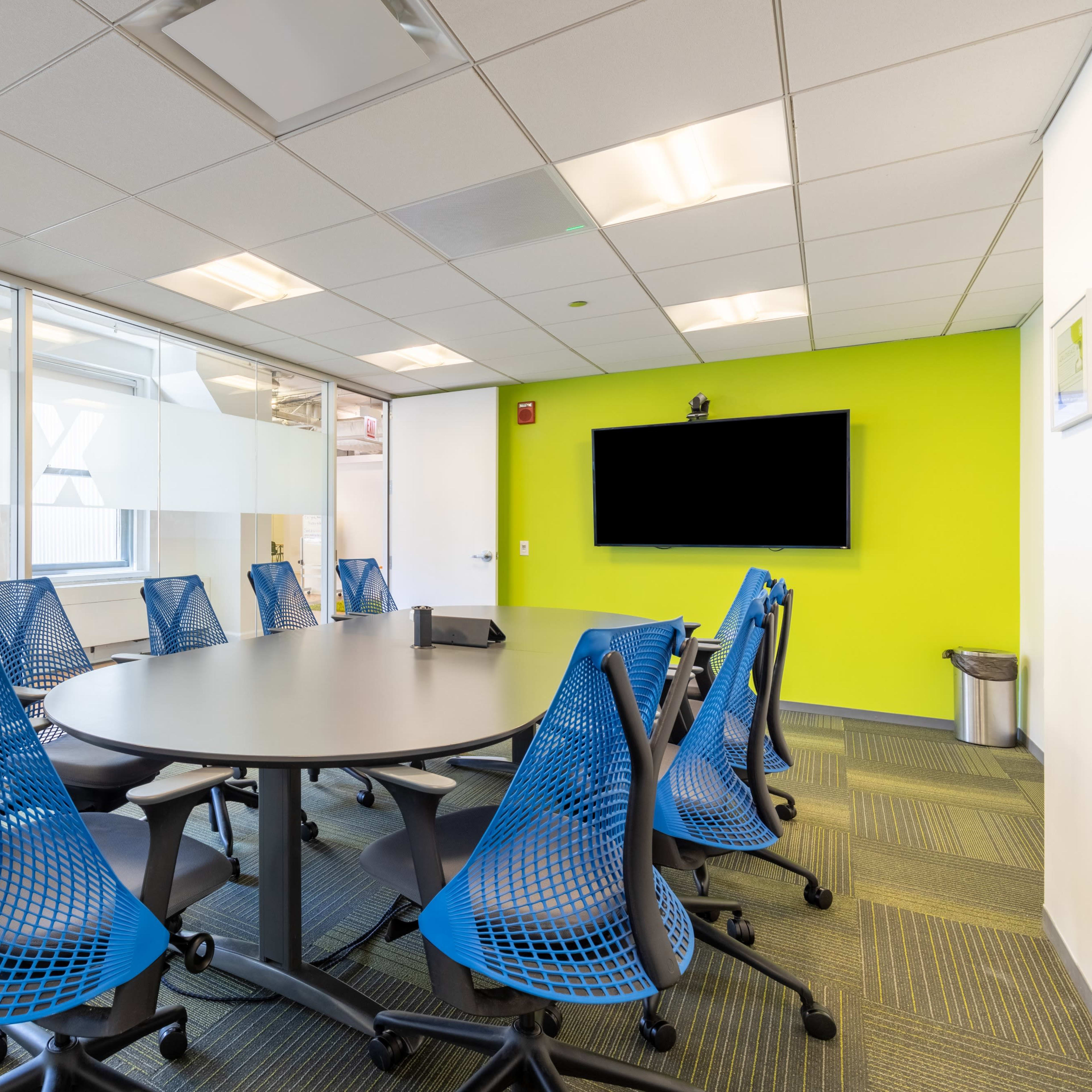 A modern conference room features a round table surrounded by blue mesh chairs, glass walls, and a large television screen mounted on a green accent wall.