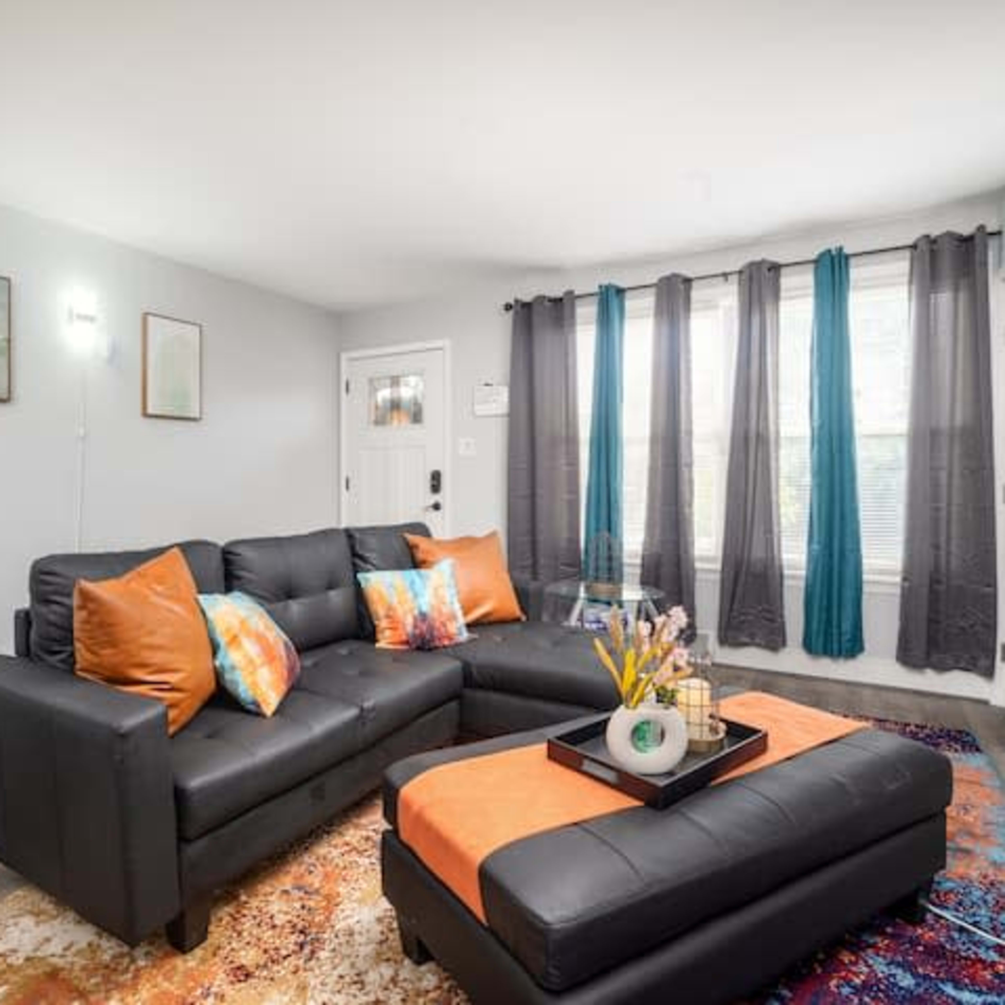 A modern living room featuring a black sectional sofa with colorful pillows, a coffee table, and large windows dressed with dark curtains.