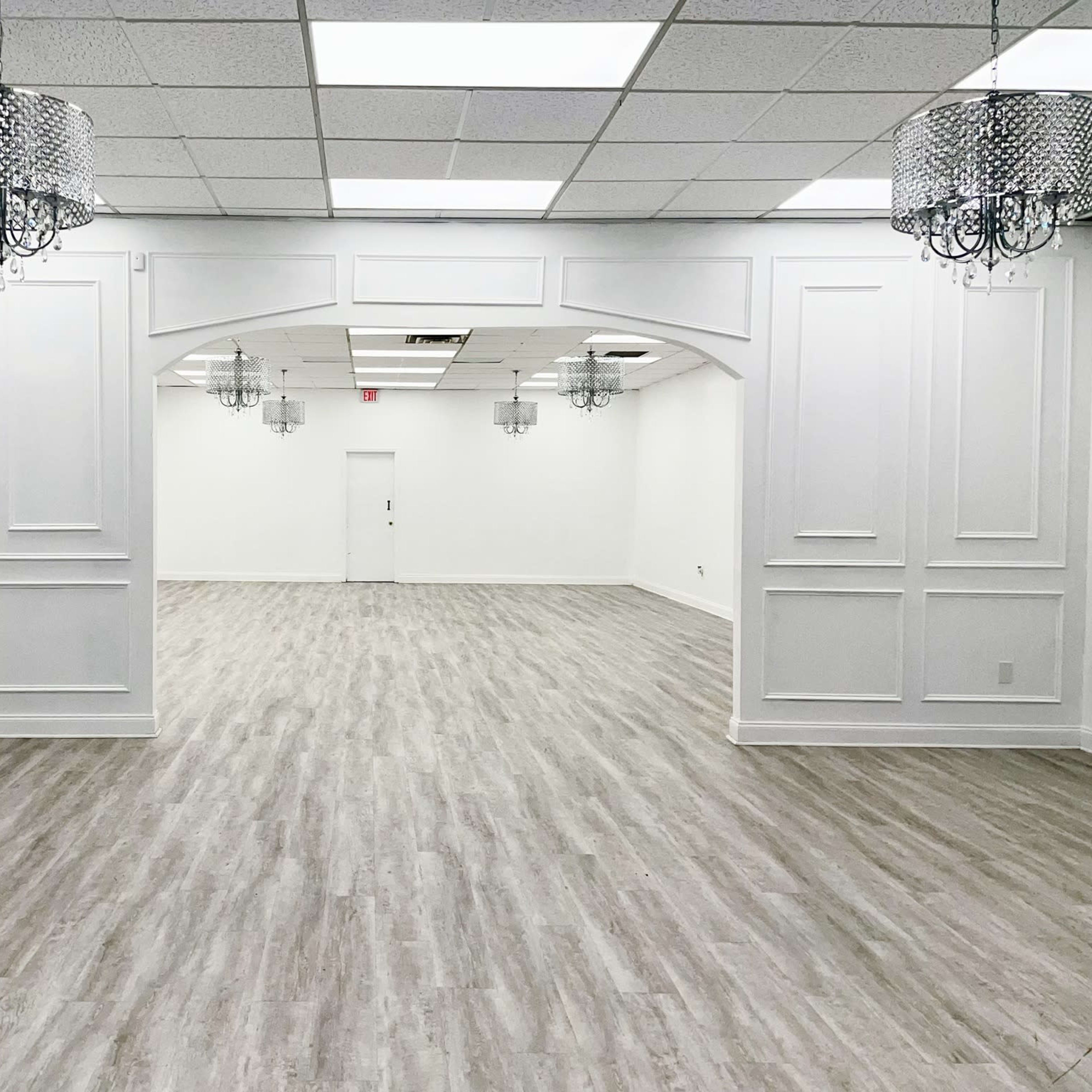 A large, empty room with white walls and a light wooden floor, featuring several decorative chandeliers hanging from the ceiling.