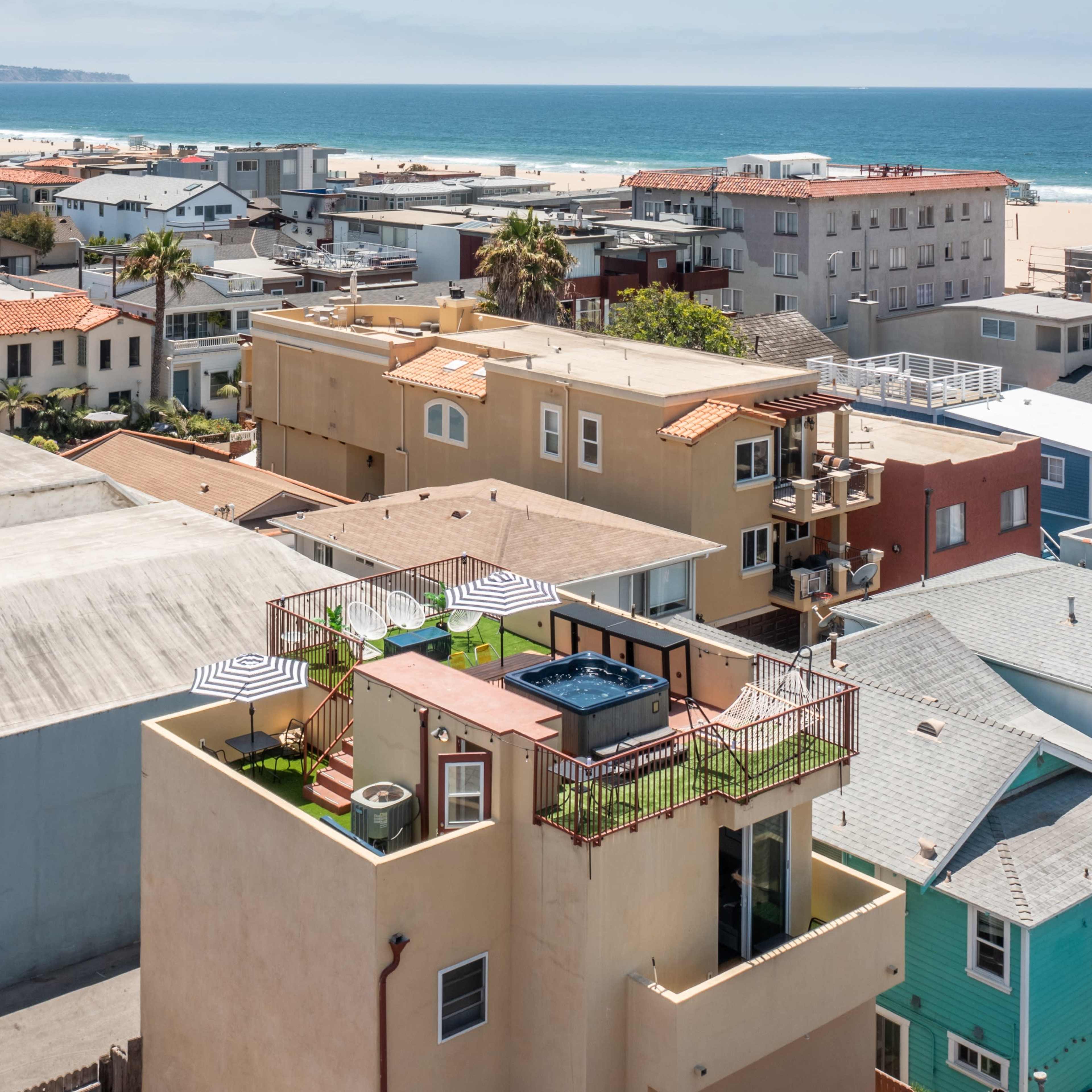 A rooftop deck with a hot tub and lounge chairs is situated atop a multi-story building among a cluster of houses near a beach.