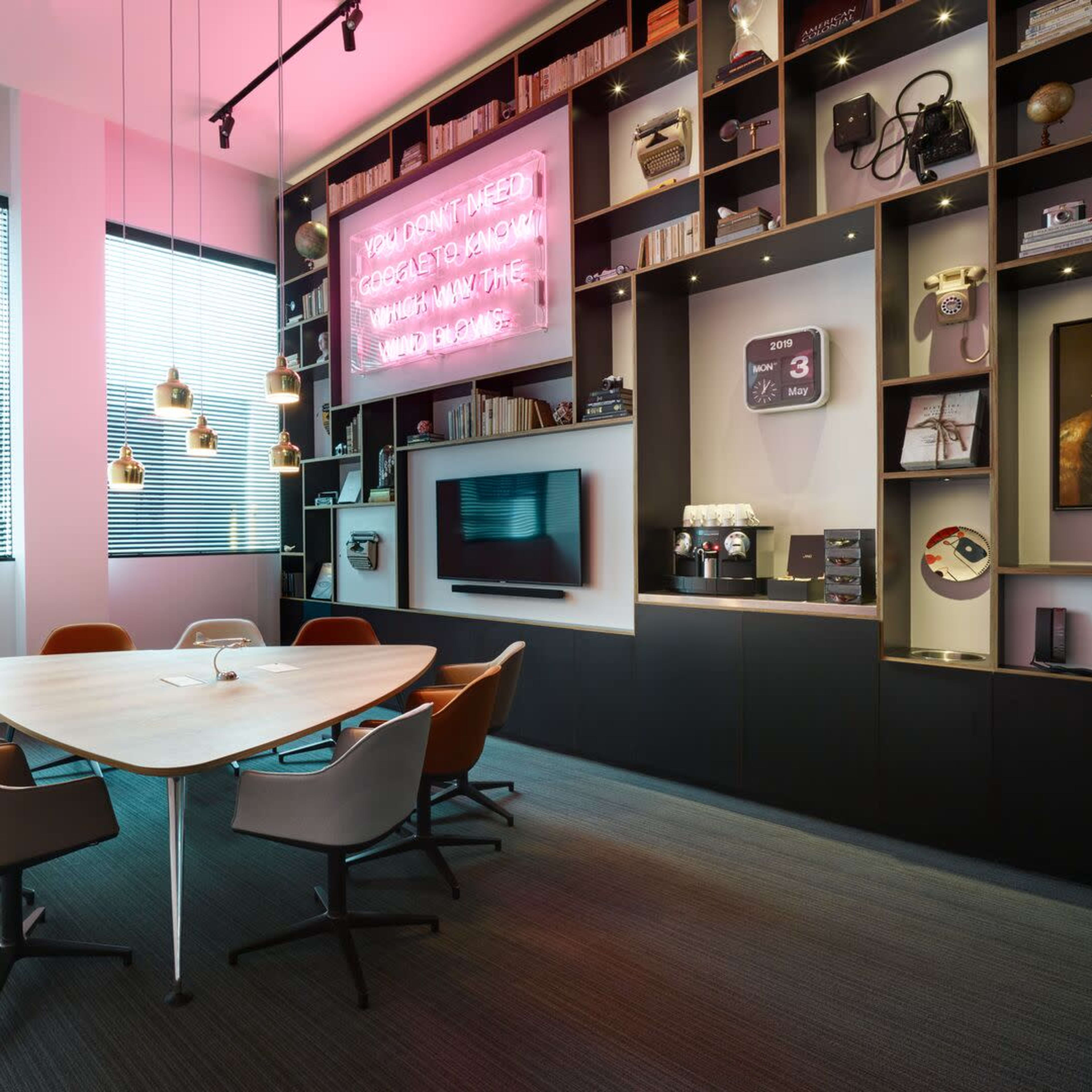 A modern conference room features a round wooden table surrounded by six chairs, with a bookshelf displaying decor and a neon sign on the wall.