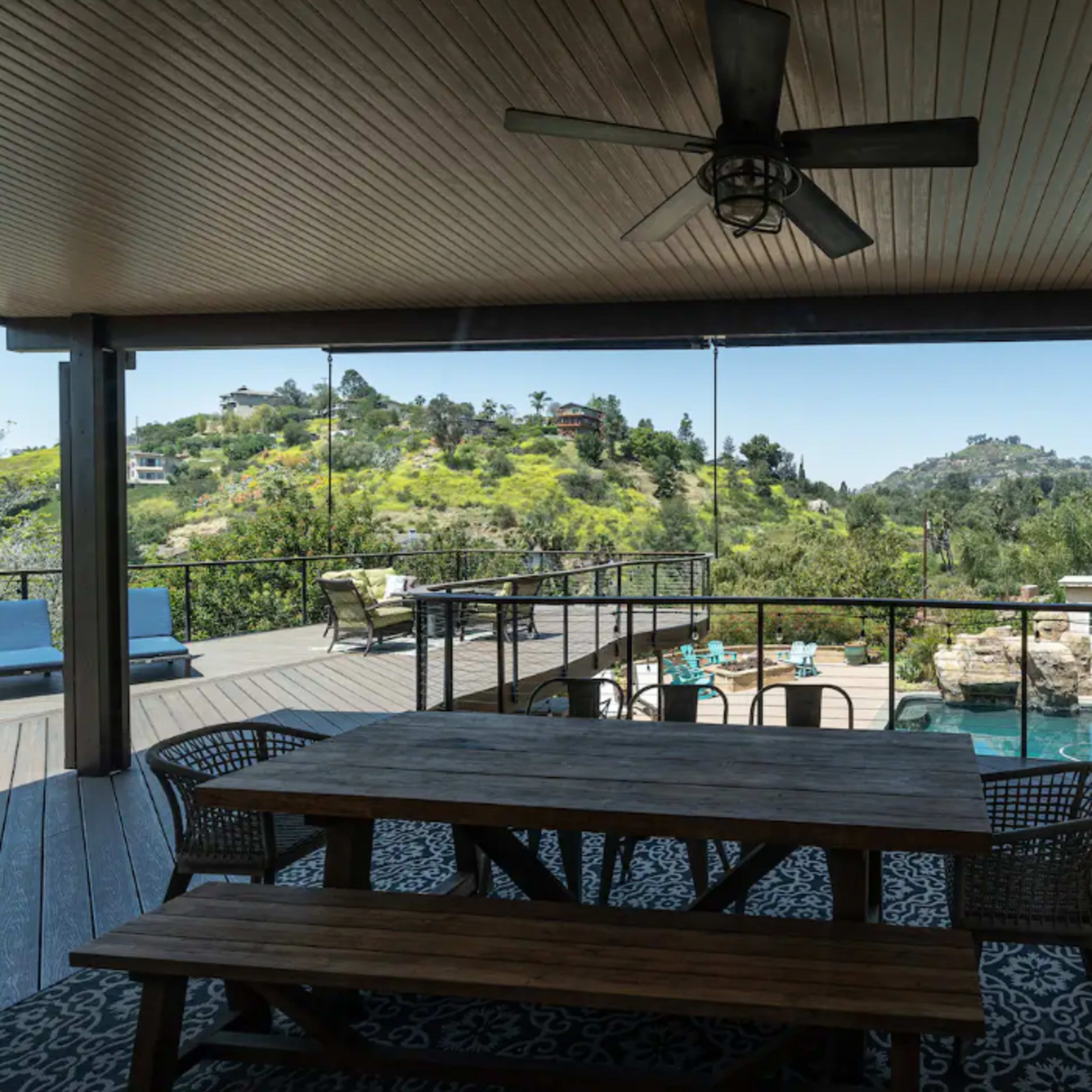 A covered patio with a wooden dining table overlooks a pool area and landscaped hills.