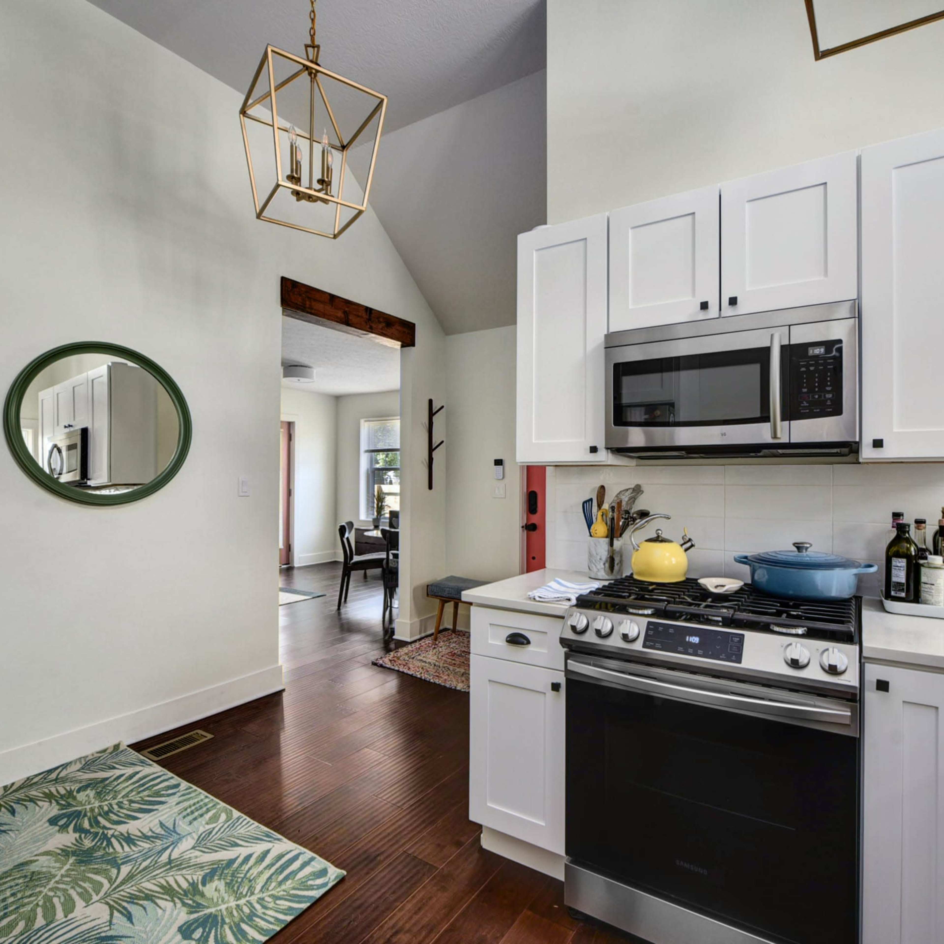 A modern kitchen with white cabinets, stainless steel appliances, and a round mirror on the wall, opens into a well-lit living space.