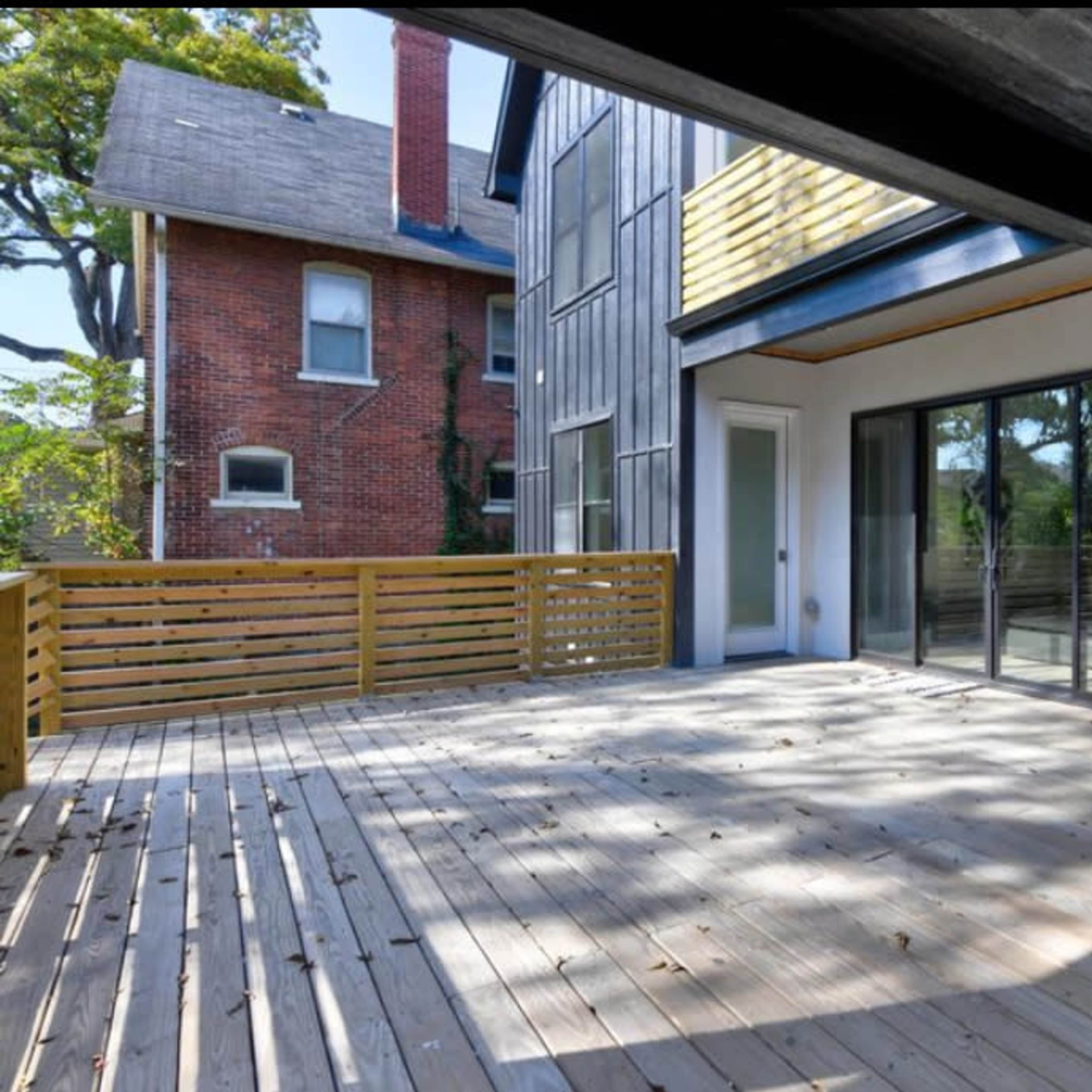 An empty wooden deck area with large glass doors leading into a modern interior space, surrounded by greenery and adjacent brick and contemporary buildings.